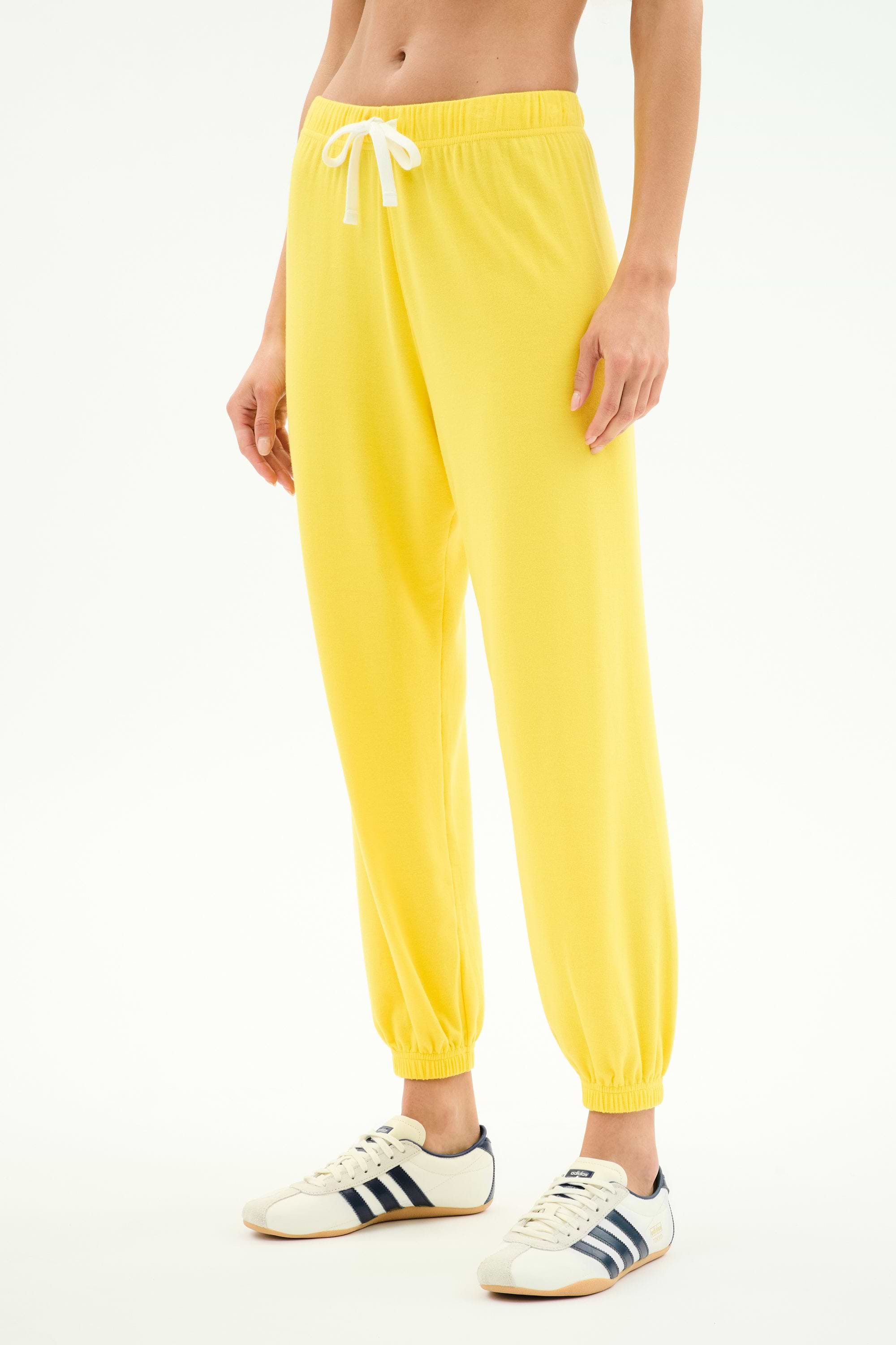 A person wears the SPLITS59 Andie Oversized Fleece Sweatpant in Bright Yellow, featuring a soft spandex blend and white drawstring, paired with white sneakers with dark stripes, standing against a plain white background.
