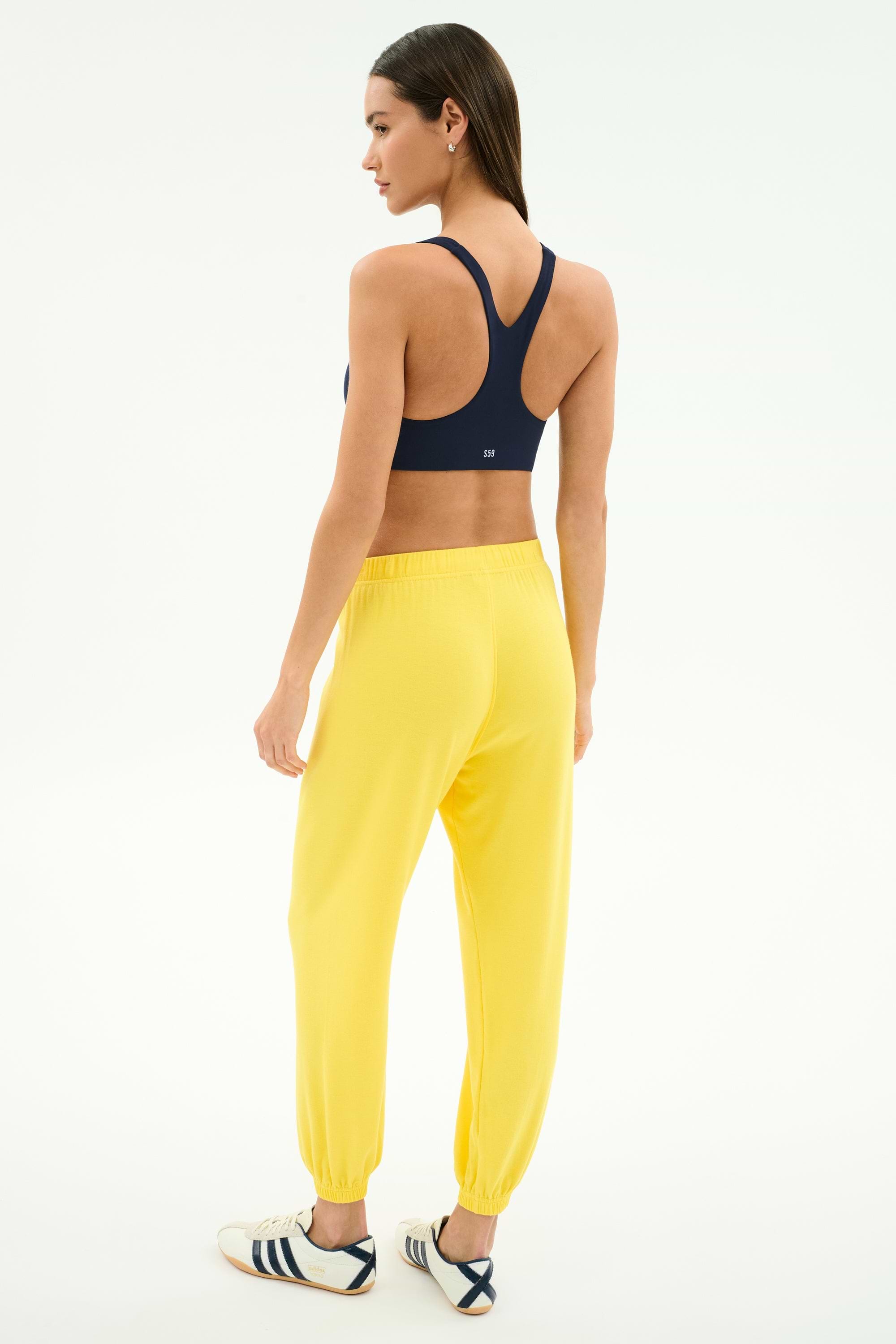 A woman faces away, wearing SPLITS59's Andie Oversized Fleece Sweatpant in bright yellow and a navy sports bra, paired with white athletic shoes with navy stripes, all set against a plain white background.