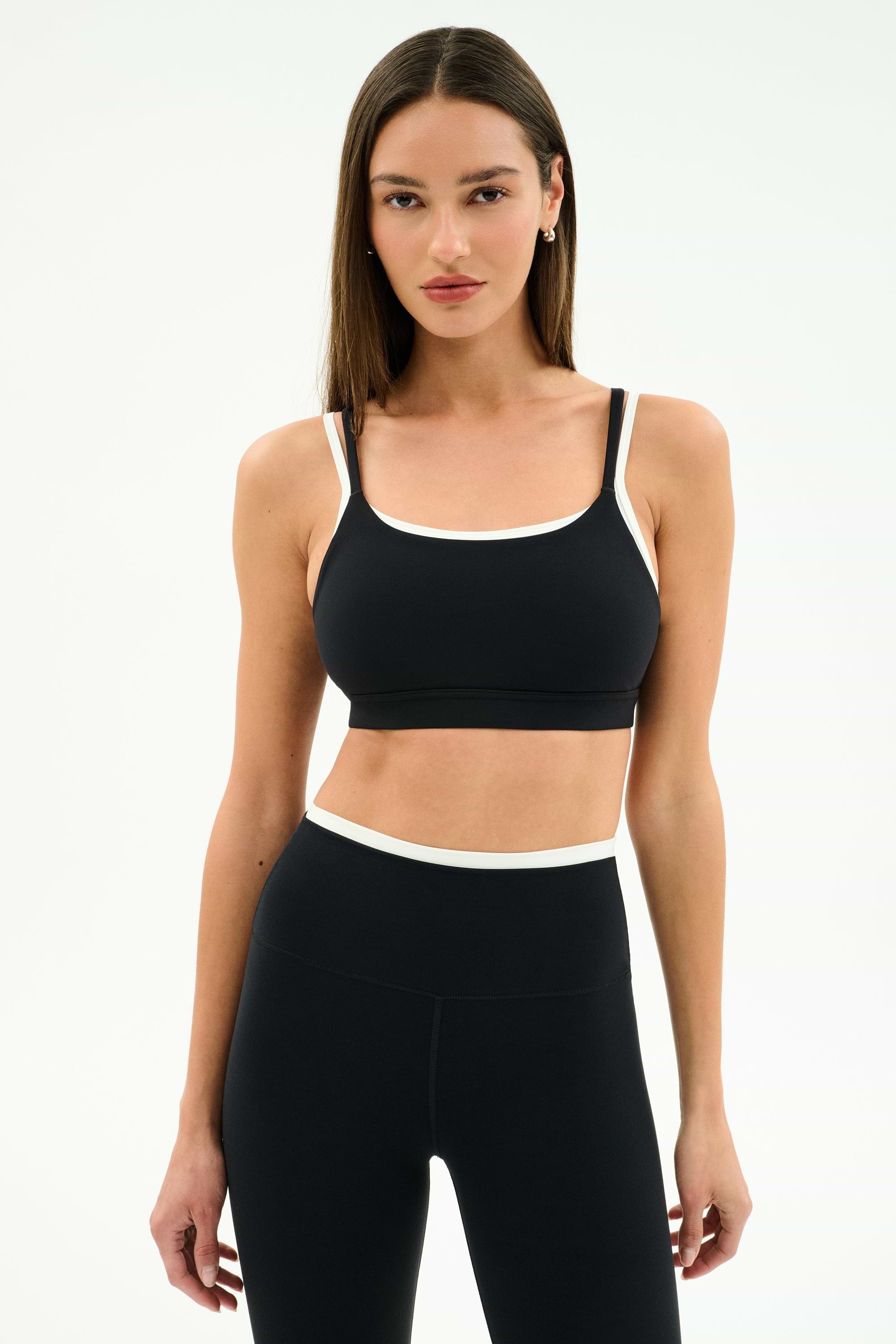 A woman wears the SPLITS59 Elliah Airweight Double Layer Bra in Black/White and high-waisted black nylon leggings, posing against a plain white background.