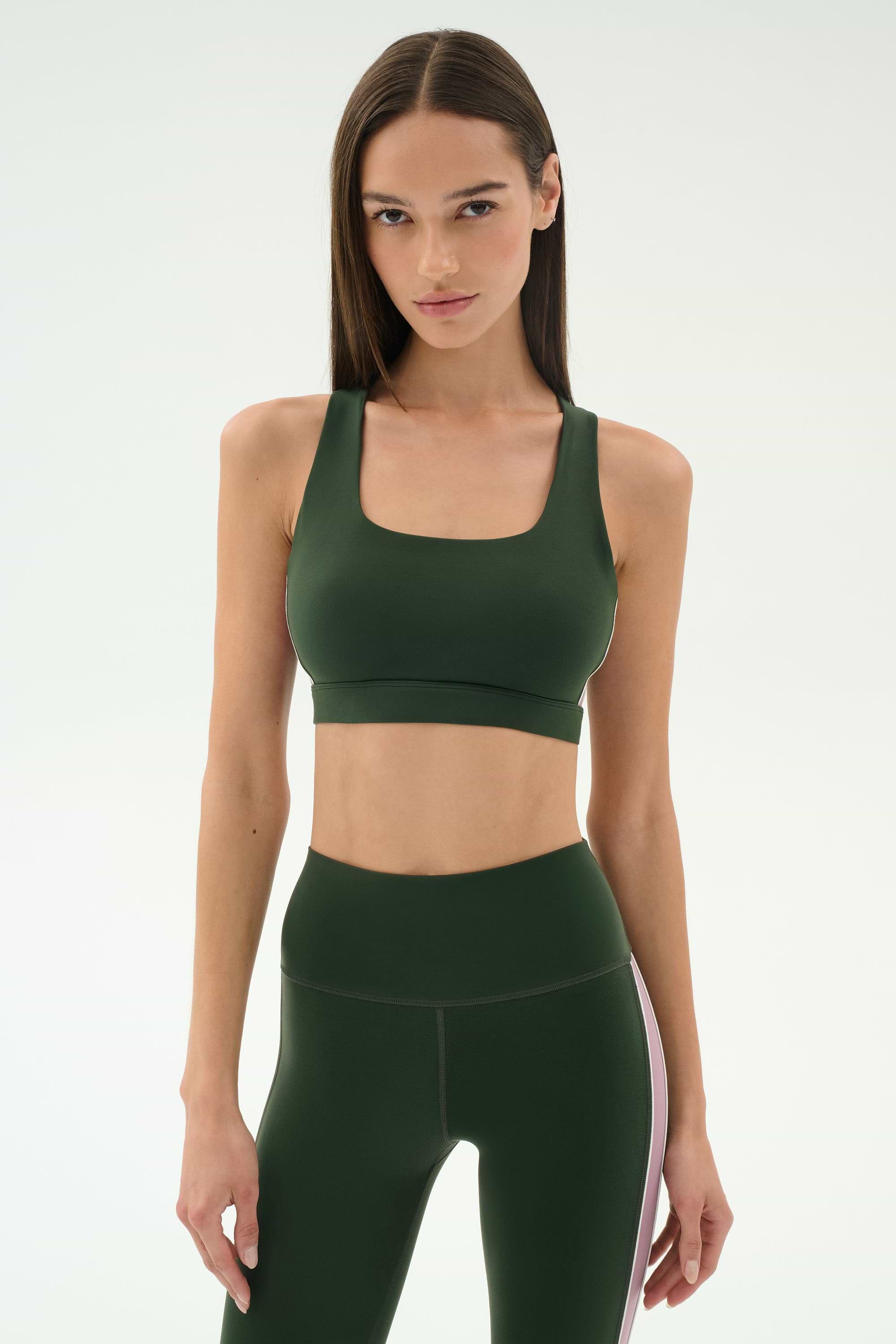 A woman faces forward, wearing the SPLITS59 Lexi Rigor Bra in Military/Blush and matching high-waisted leggings with a white side stripe, set against a plain light background.