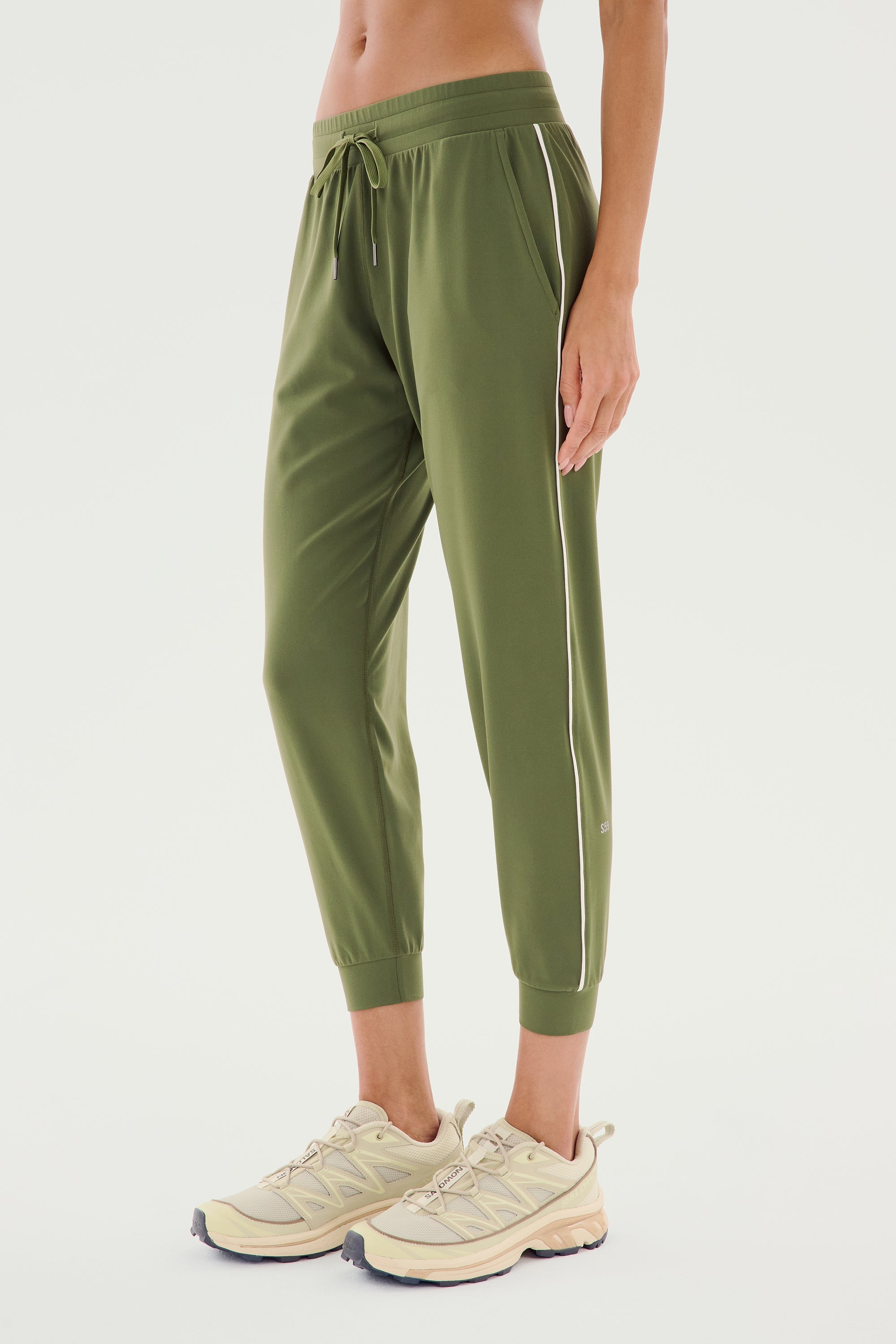 A person is standing, showcasing the lower half of their body. They are wearing SPLITS59's Airweight Jogger 7/8 with Piping in Olive/White and a pair of beige sneakers. The joggers feature a touch of spandex fabric for enhanced comfort. The background is plain white.