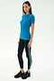 A woman stands against a plain backdrop wearing the SPLITS59 Louise Rib Short Sleeve in Stone Blue, paired with black leggings featuring side stripes and black sneakers—ideal for gym workouts or yoga.