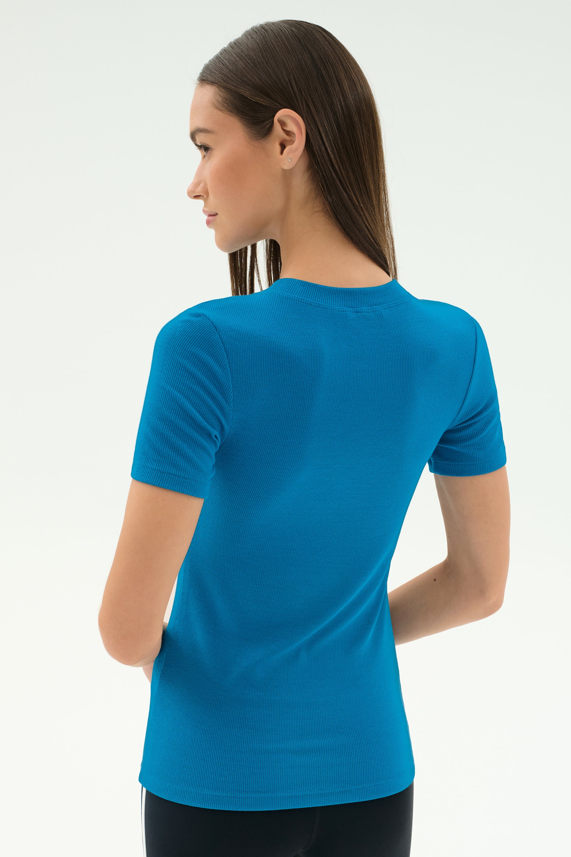 A woman with straight brown hair stands with her back to the camera, wearing the SPLITS59 Louise Rib Short Sleeve in Stone Blue against a plain light background.