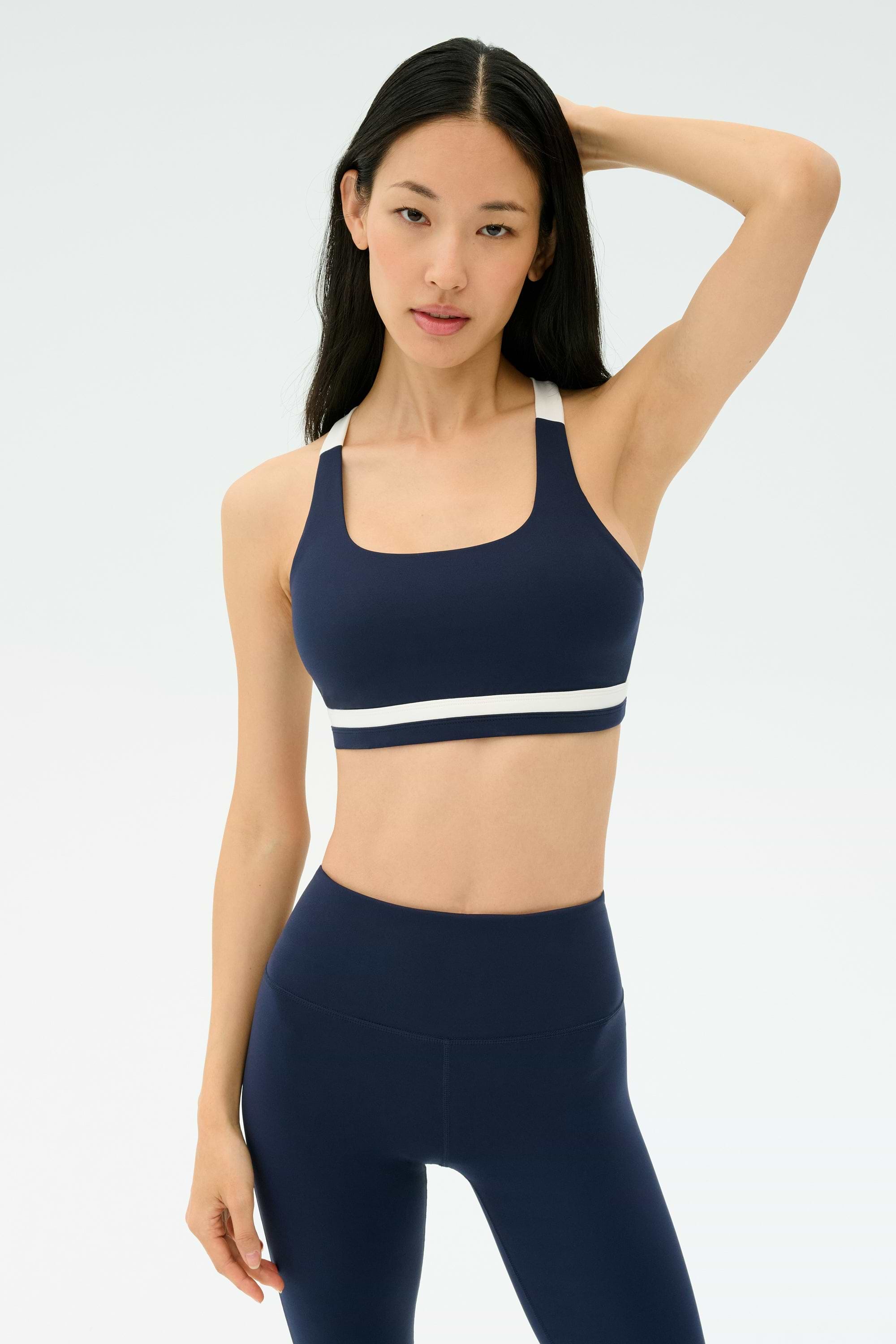 A woman wears the SPLITS59 Miles Rigor Bra in Indigo/White, standing against a plain light background with one hand on her head and a neutral expression.