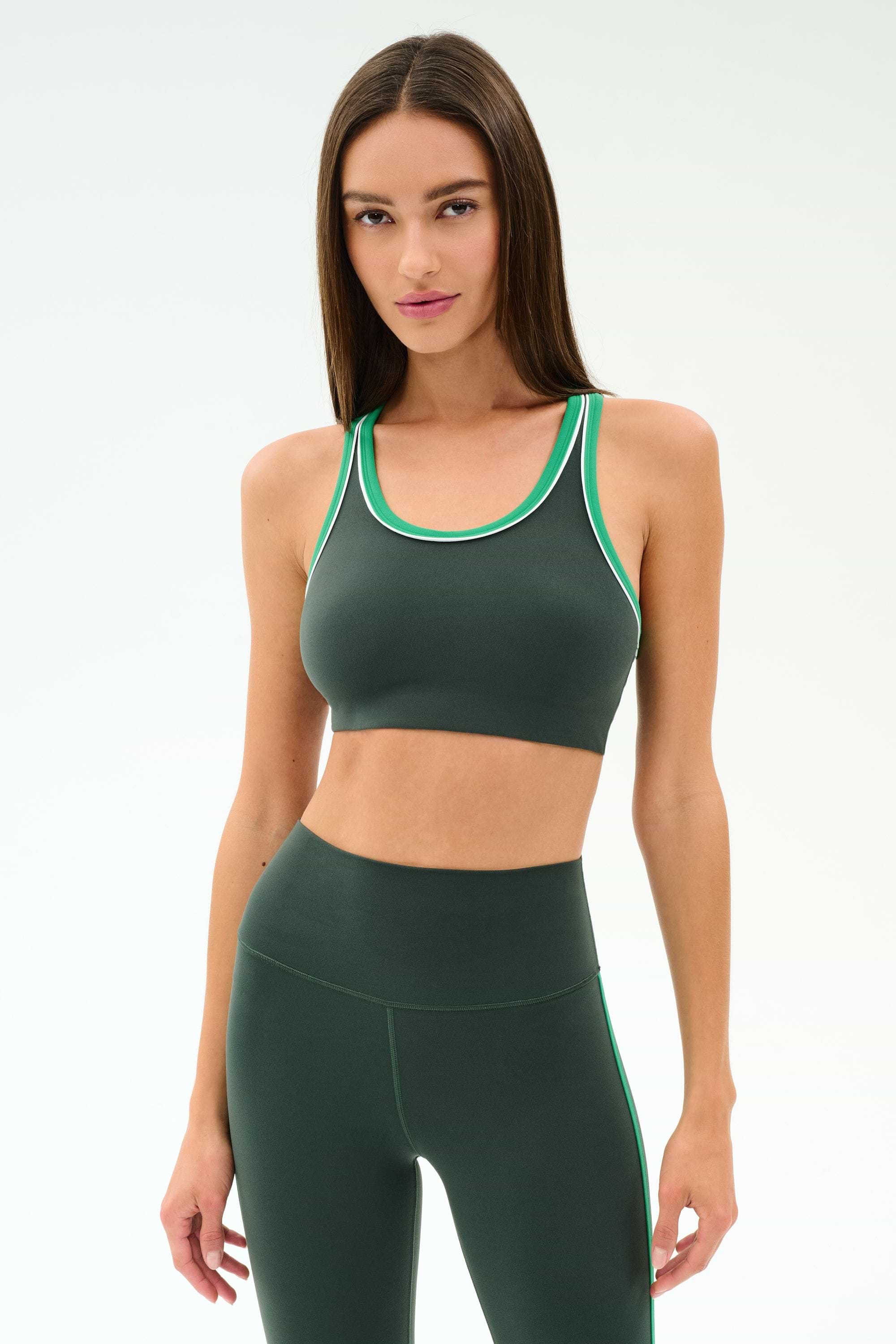 A woman wears the SPLITS59 Robbie Airweight Bra in Military/Spearmint, featuring dark green with light green trim, against a plain light background.