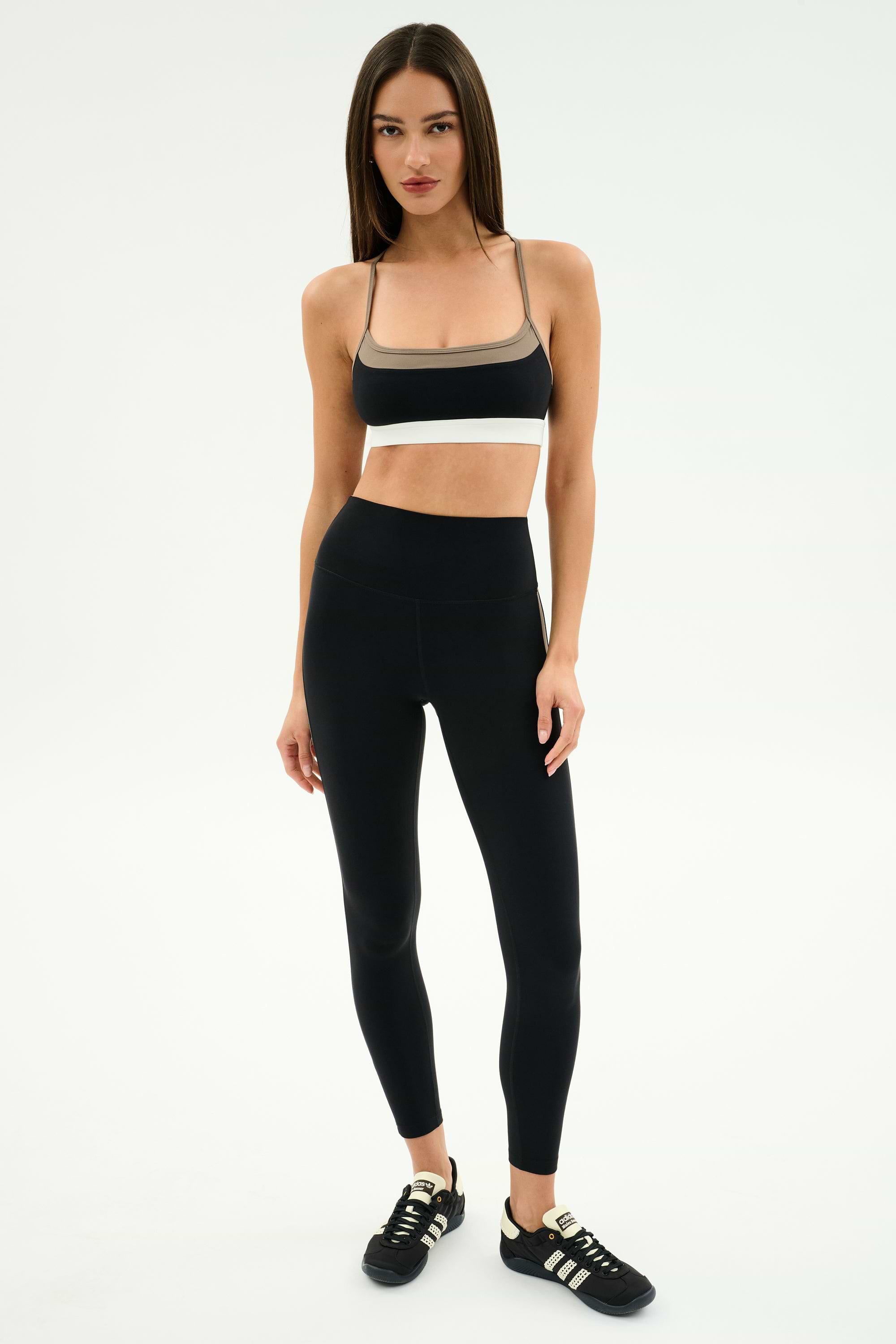 A woman stands against a plain background, wearing the SPLITS59 Scottie Airweight Bra in Black/Lentil, paired with high-waisted black leggings and athletic shoes.