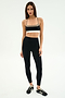 A woman stands against a plain background, wearing the SPLITS59 Scottie Airweight Bra in Black/Lentil, paired with high-waisted black leggings and athletic shoes.