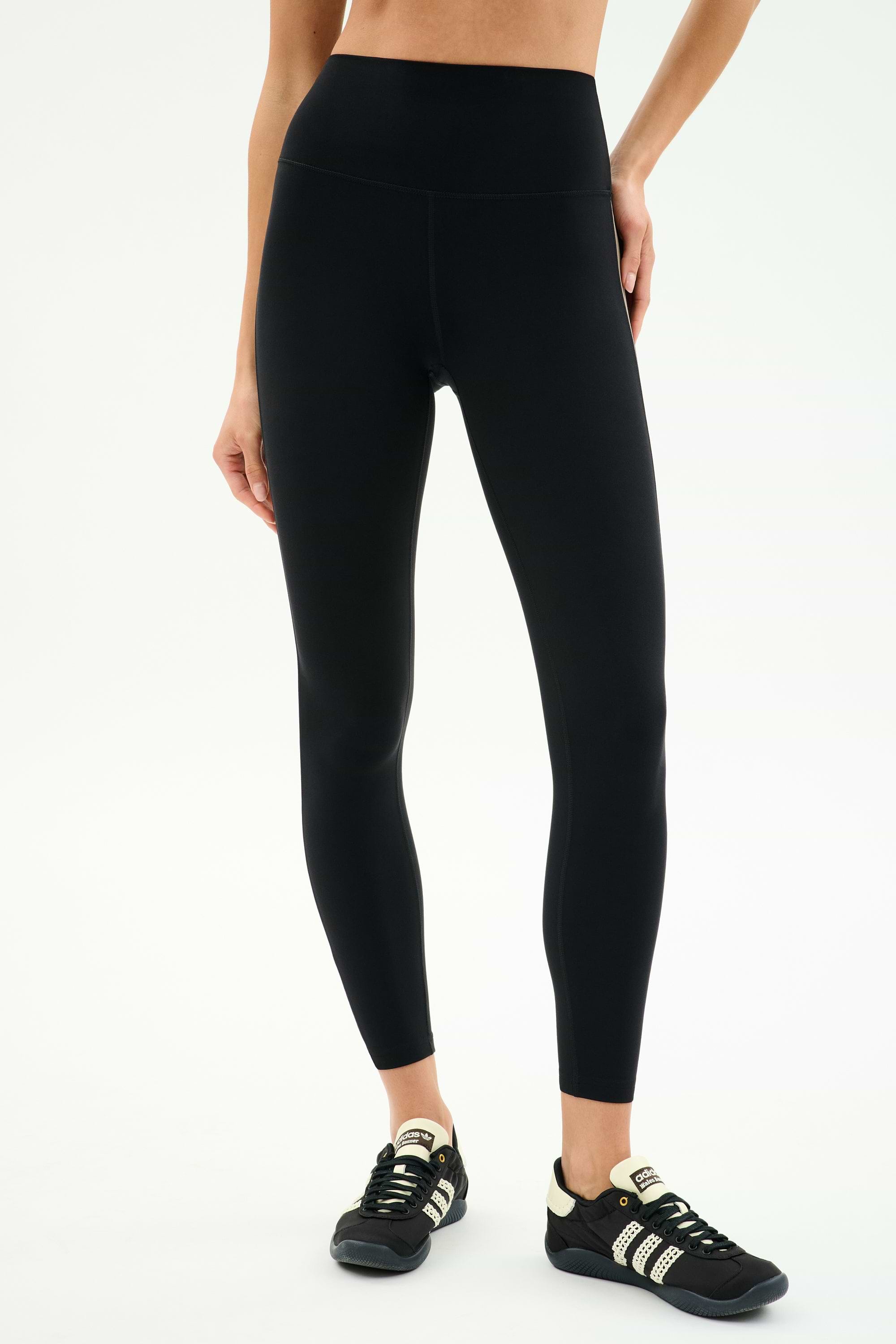 Wearing SPLITS59 Layla Airweight High Waist 7/8 in Black/Lentil and black sneakers with white stripes, standing against a light background—ideal for hot yoga or any workout.
