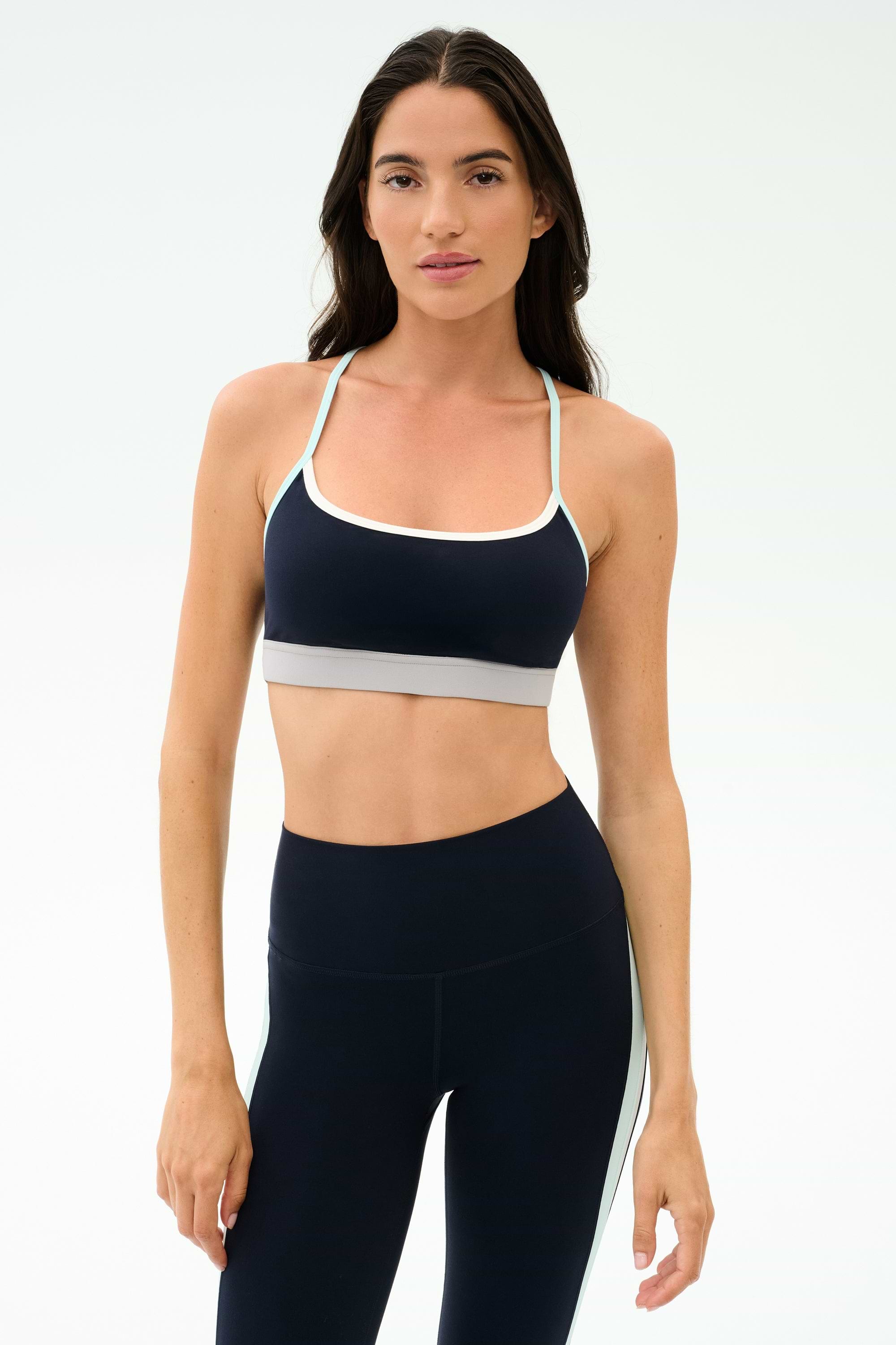A woman stands against a plain background wearing the SPLITS59 Emerson Airweight Bra in Indigo/Dove Grey, paired with matching high-waisted leggings—ideal for Pilates or hot yoga.