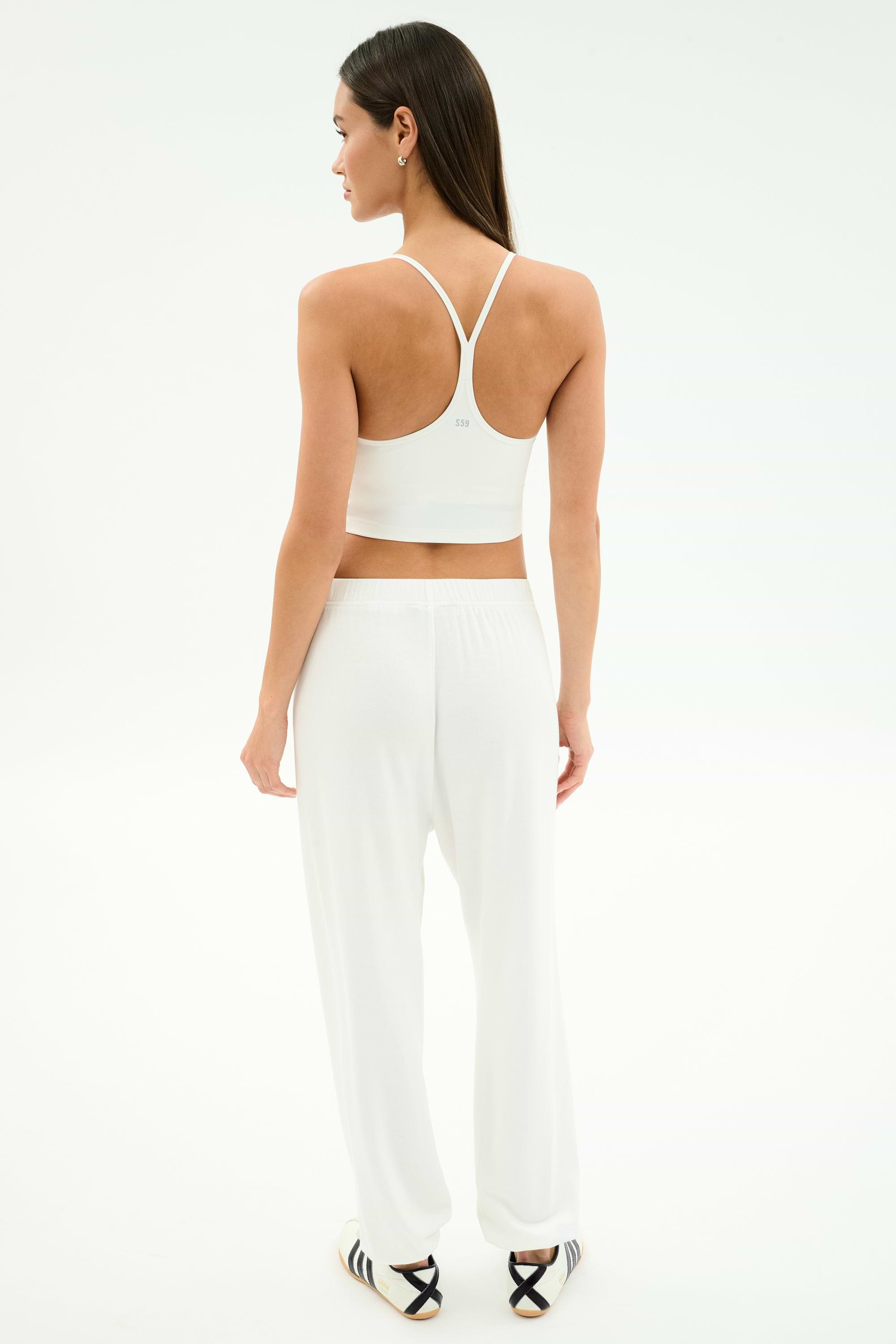 A woman faces away, wearing SPLITS59's Andie Oversized Fleece Varsity 59 Sweatpant in White/59 with a white sports bra and black-striped slide sandals, perfect for cool weather workouts, set against a plain white background.