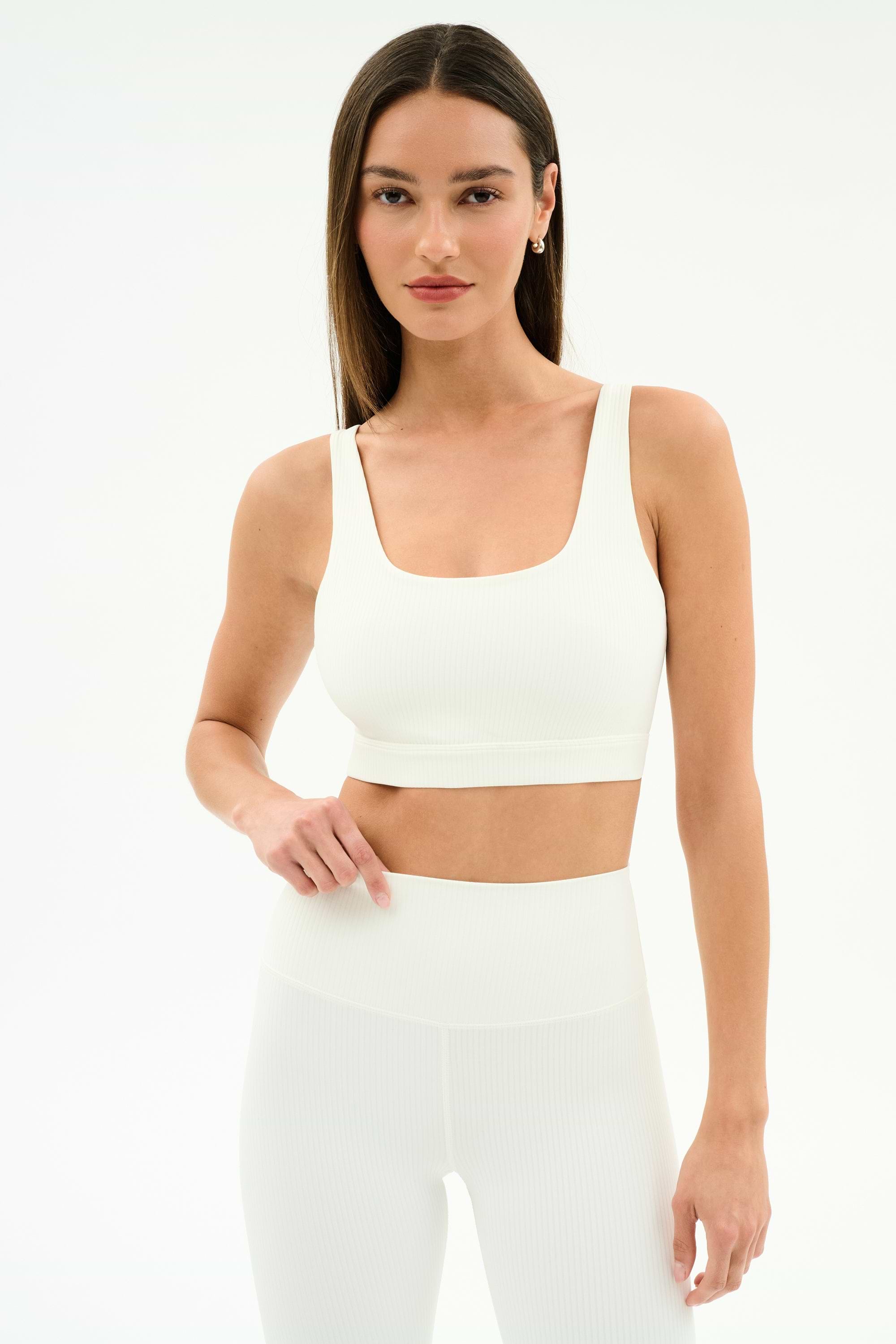 A woman wears the SPLITS59 Airweight Rib Bra in white, crafted from moisture-wicking ribbed fabric—ideal for hot yoga—paired with matching high-waisted leggings, standing against a plain white background.