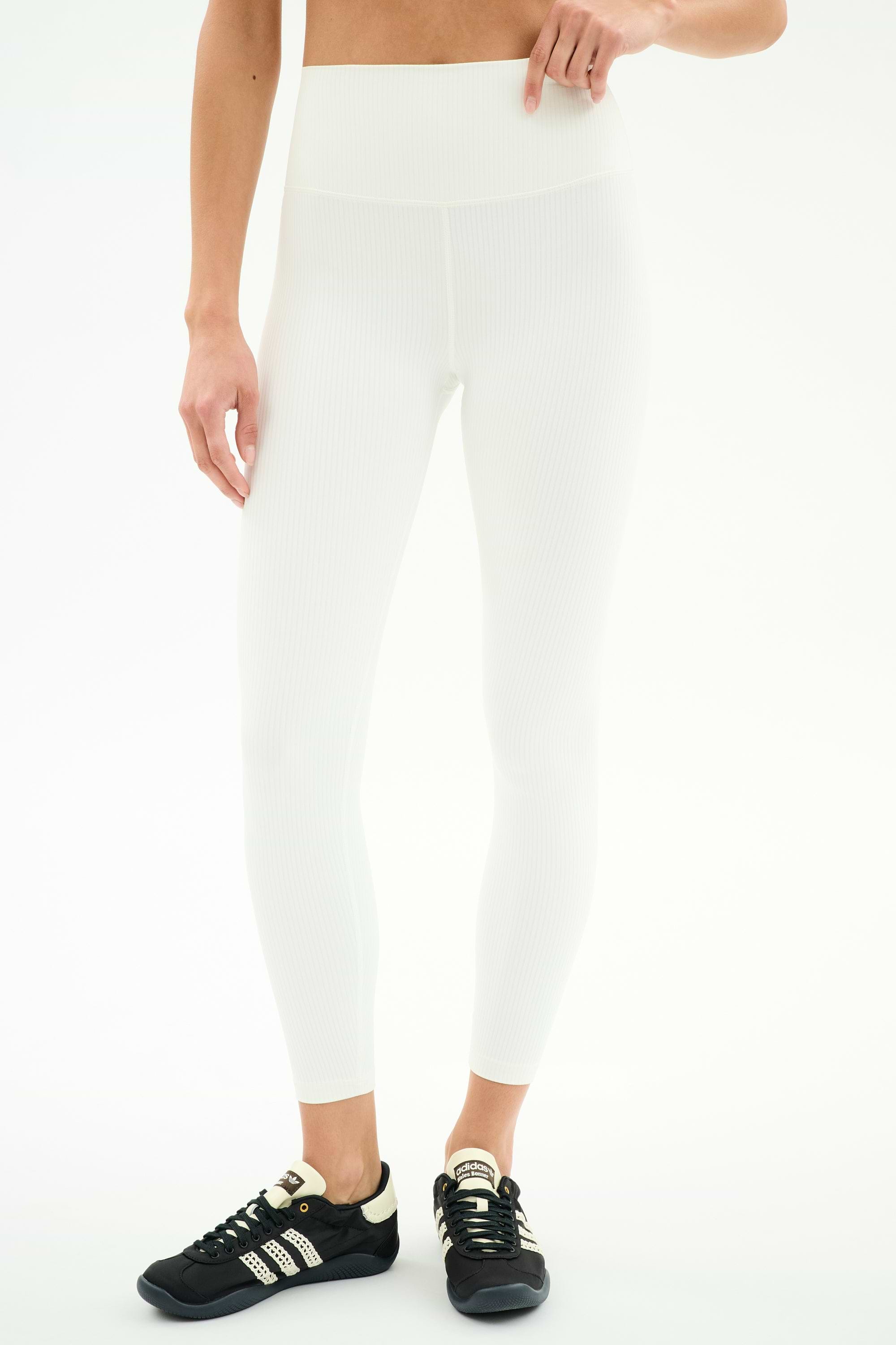 Person wears SPLITS59 Airweight Rib High Waist 7/8 leggings in white with black Adidas sneakers, standing against a plain white background. Upper body partially out of frame—ideal for Pilates or hot yoga.