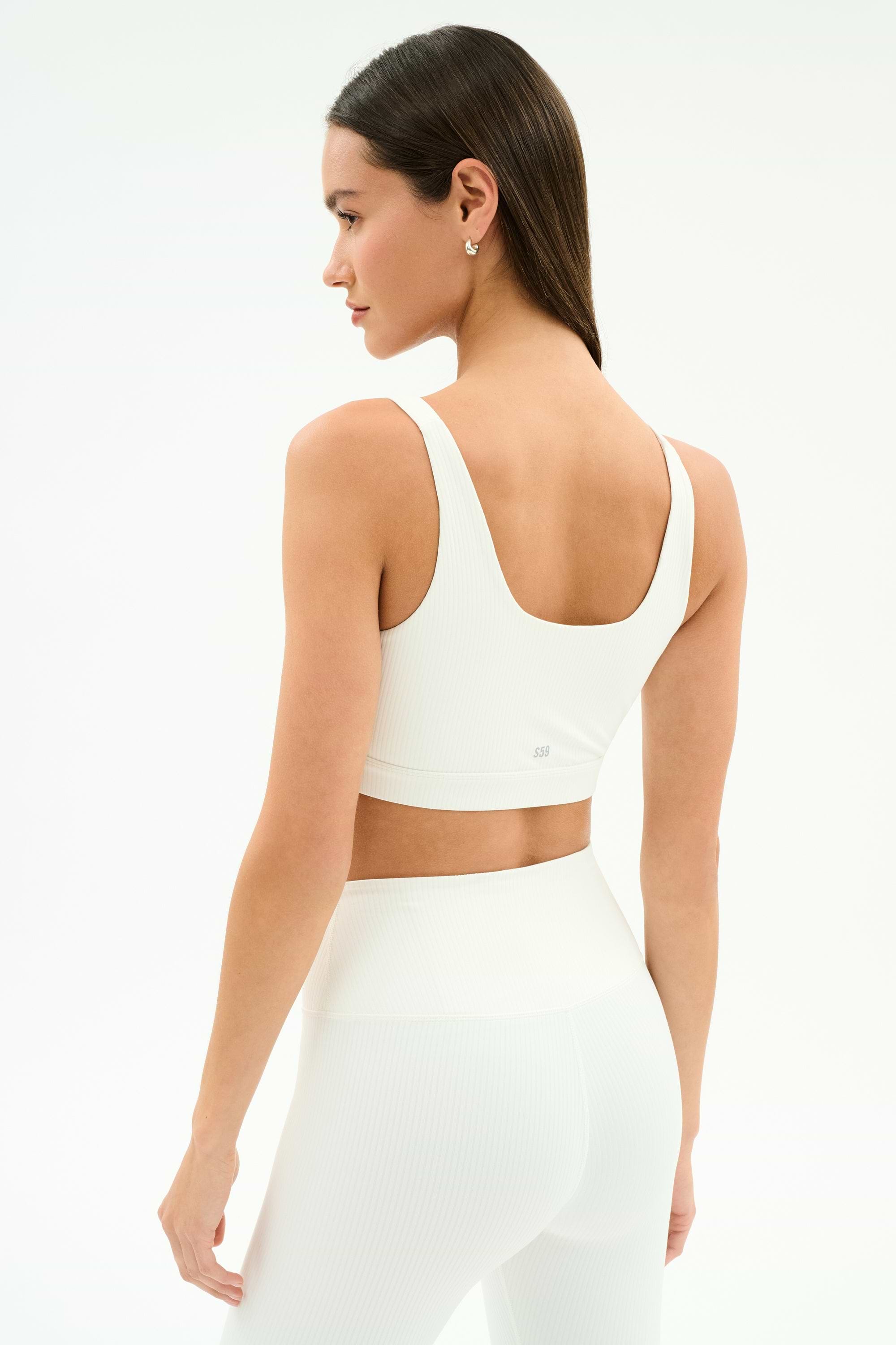 A woman models the SPLITS59 Airweight Rib Bra in white, turning to her left to showcase its breathable ribbed fabric—ideal for hot yoga—against a plain white background.