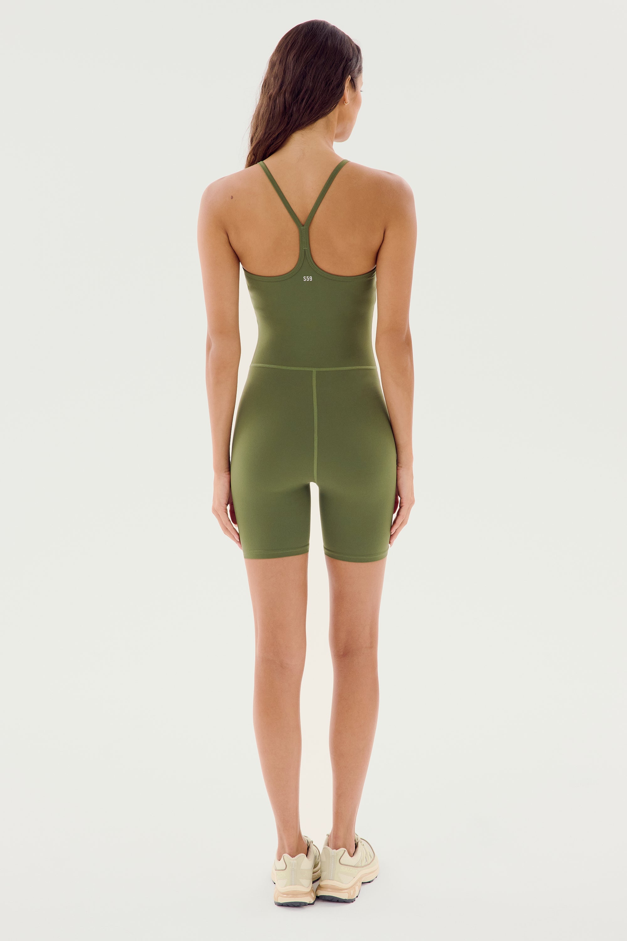 A person stands facing away, wearing the SPLITS59 Airweight 6” Short Jumpsuit in olive, complemented by beige sneakers.