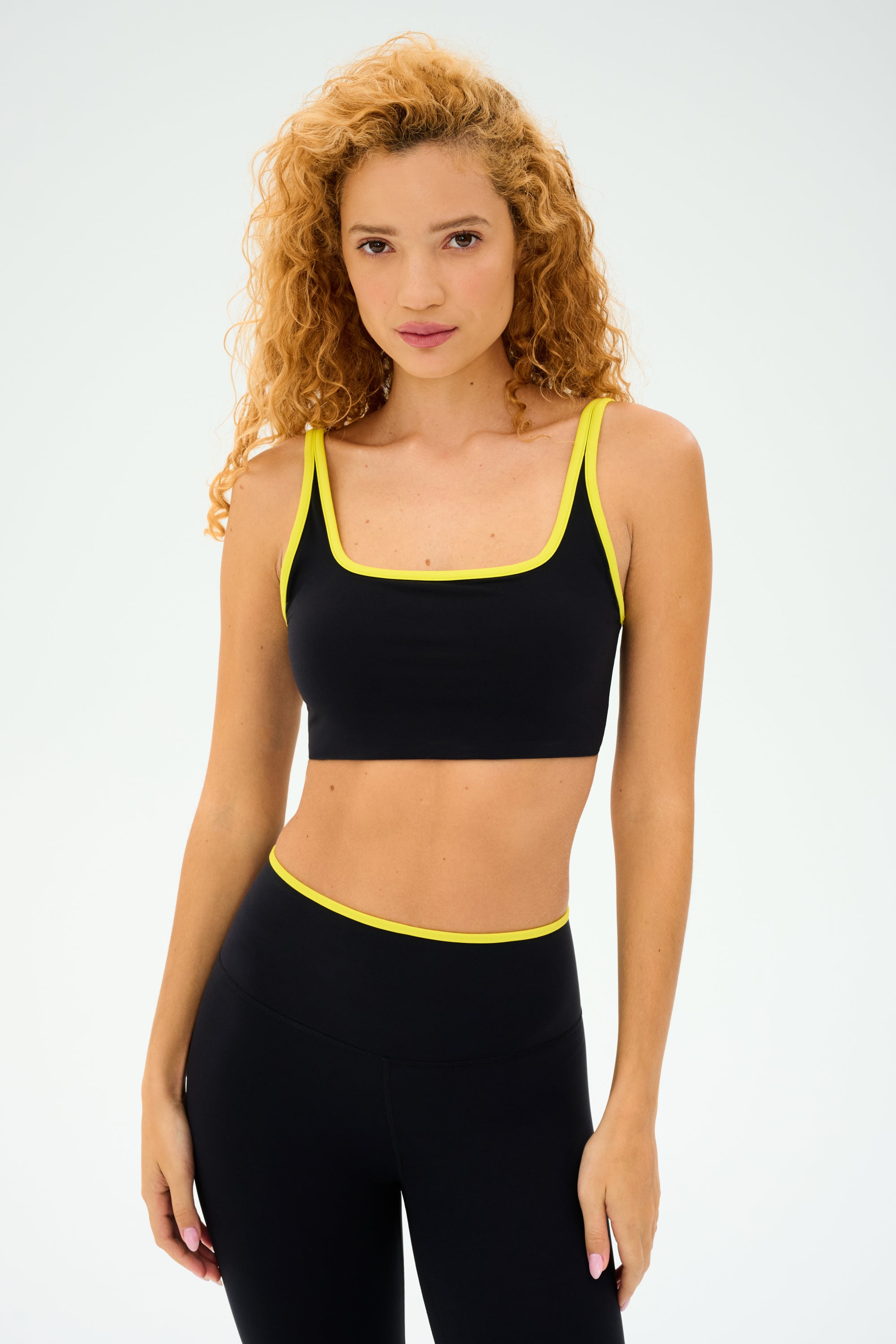 A person with curly hair confidently stands against a plain backdrop, sporting SPLITS59's Cait Rigor Bra in Black/Chartreuse, made of high-performance fabric ideal for intense workouts.