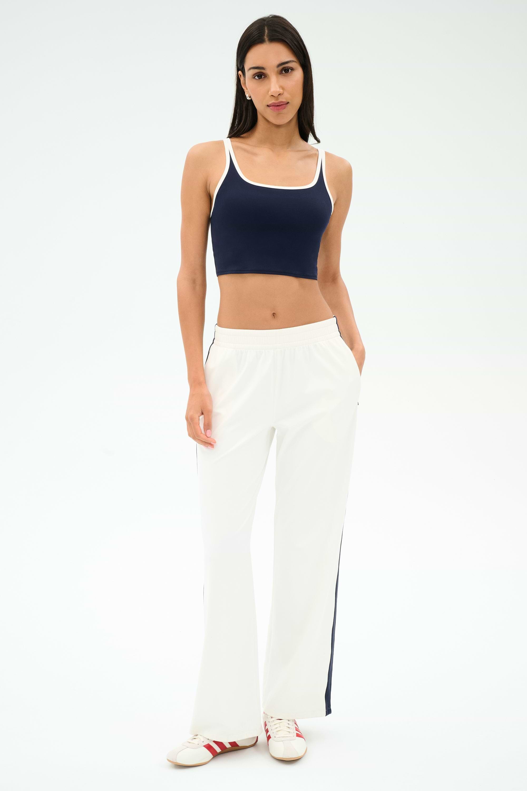 A woman stands against a plain background wearing a navy crop top, SPLITS59 Maxie Rigor Track Pant in White/Indigo with wide legs and side stripes, and white sneakers with red accents.