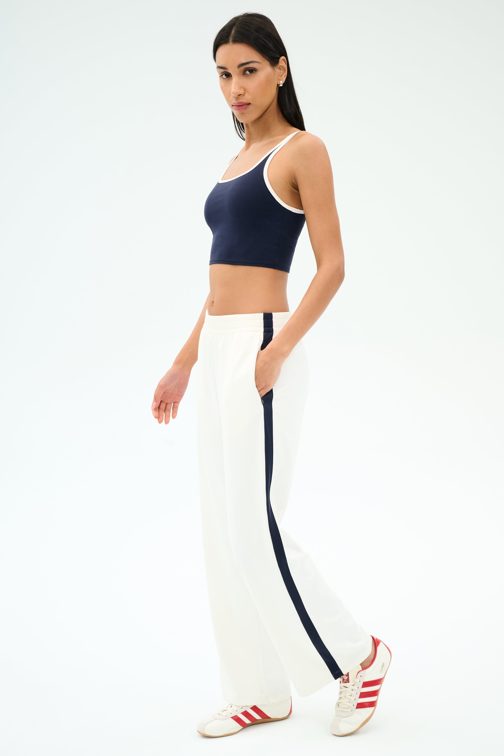 A woman poses in a navy crop top, SPLITS59 Maxie Rigor Track Pant - White/Indigo, and white sneakers with red stripes.