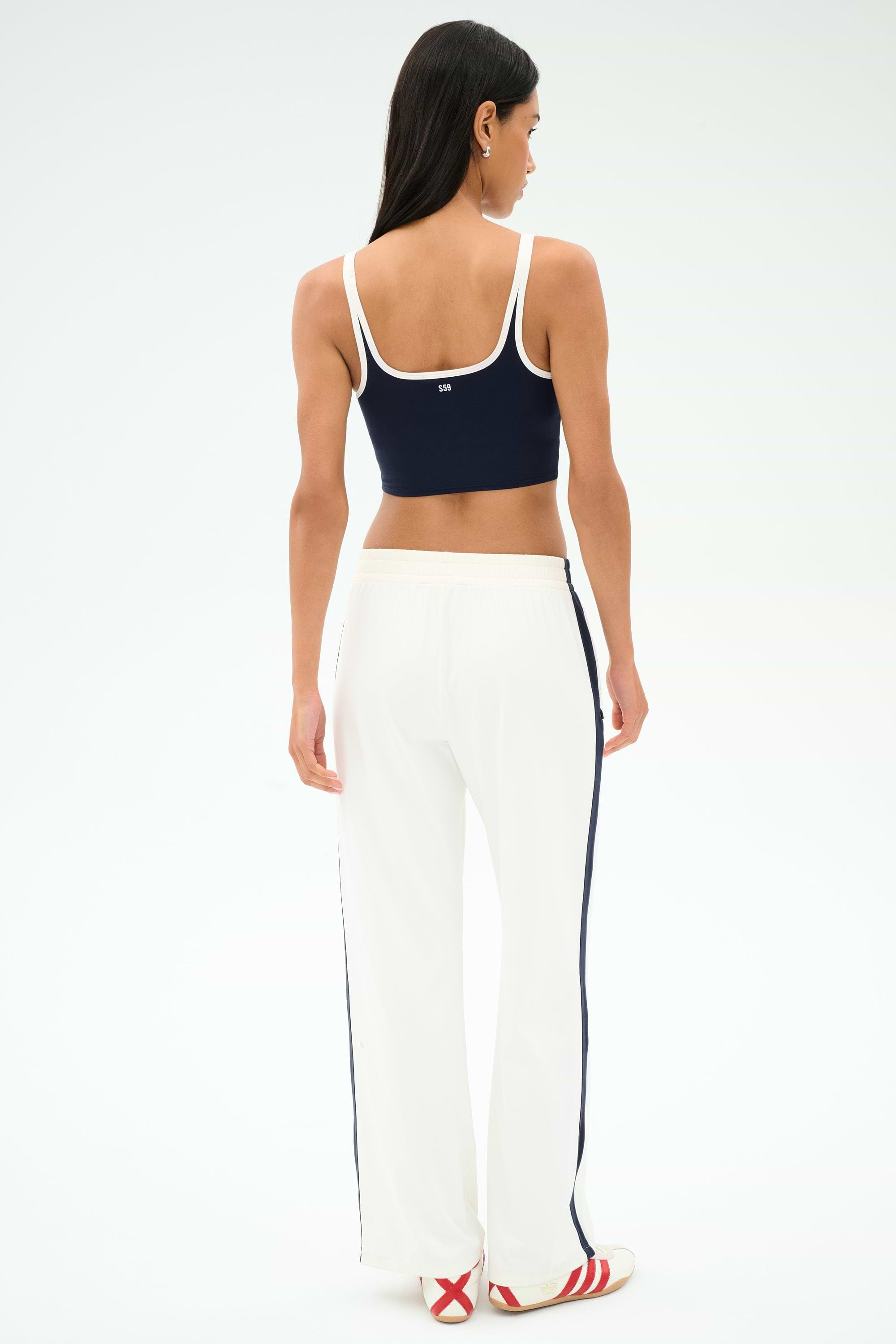 A woman faces away, wearing a navy crop top with white trim, SPLITS59 Maxie Rigor Track Pant in white/indigo featuring navy side stripes, and white sneakers with red stripes.