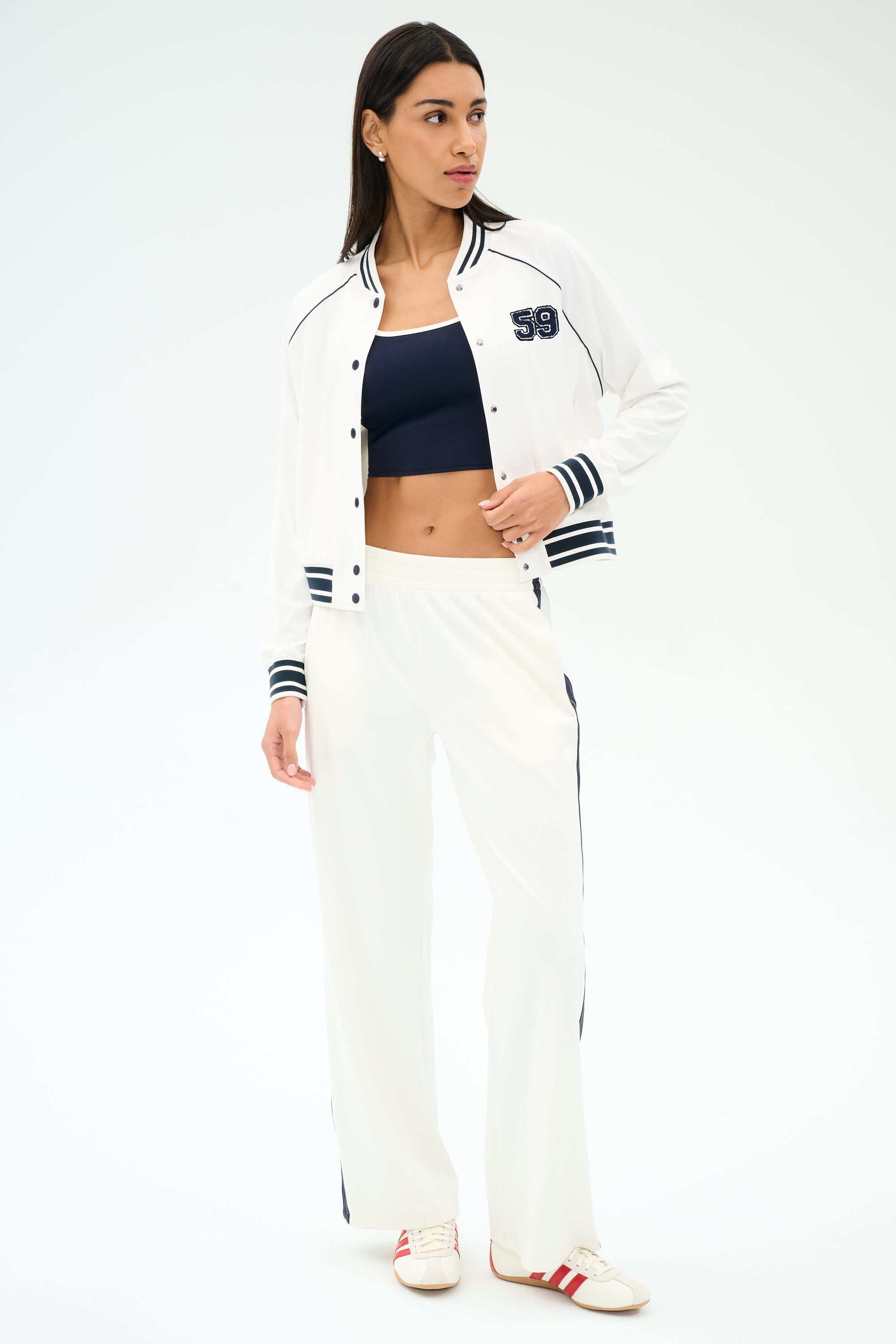 A woman wears the SPLITS59 Hayes Varsity Rigor Jacket in white, a navy crop top, spandex track pants with navy stripes, and white sneakers with red stripes.