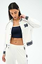 A woman wearing the SPLITS59 Hayes Varsity Rigor Jacket in white, styled over a navy sports bra and white track pants, looks down while touching her hair.
