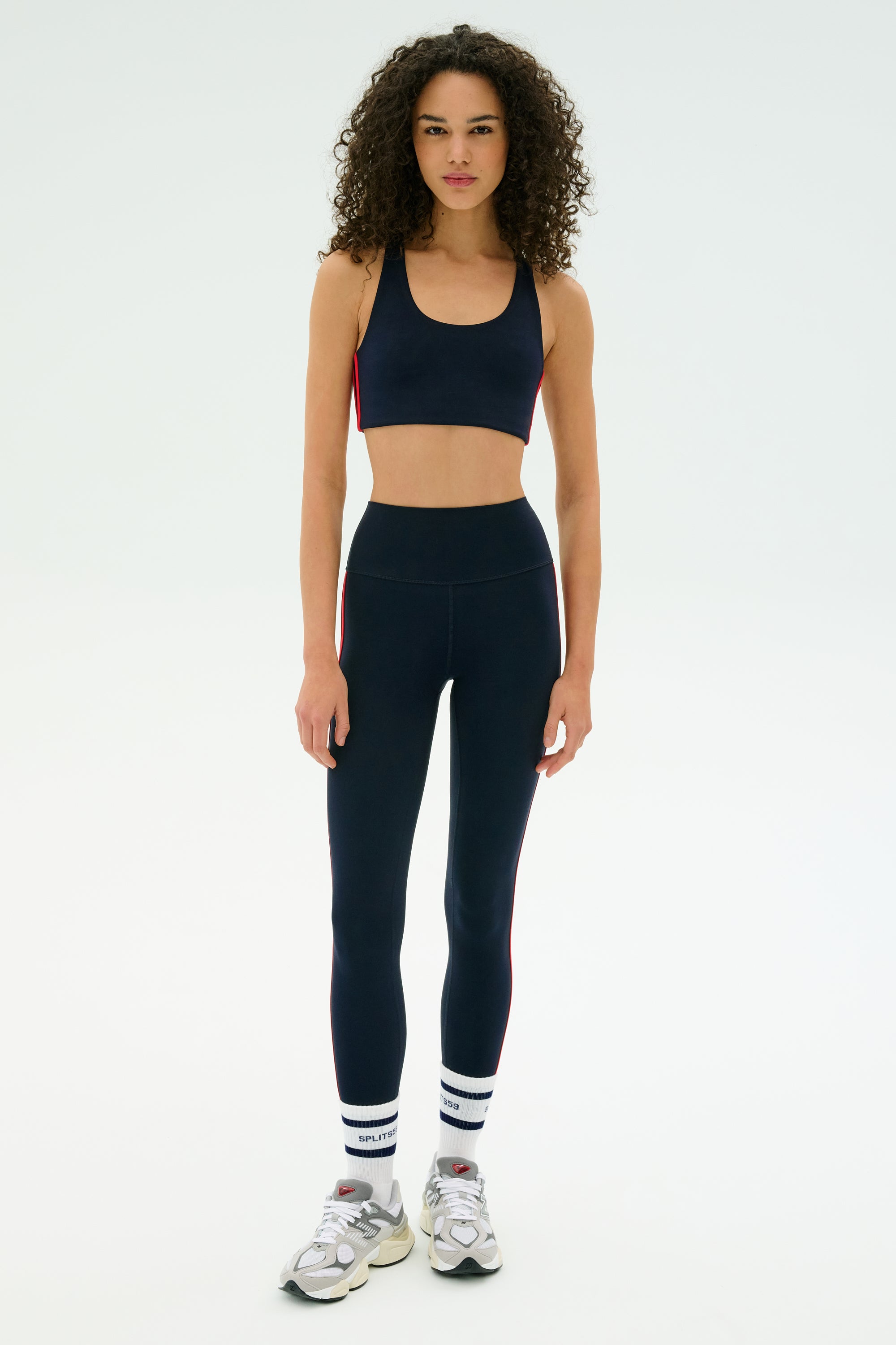 A woman faces forward in the SPLITS59 Ella Airweight Bra - Indigo/Pirate Red, paired with matching high-waisted leggings, white socks, and silver athletic shoes on a plain white background.