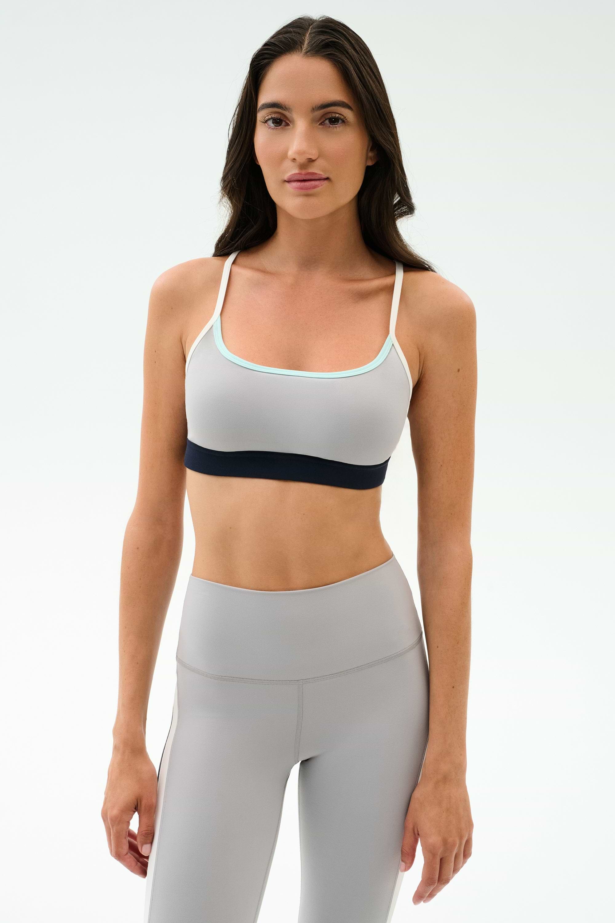 Woman wearing the SPLITS59 Emerson Airweight Bra in Dove Grey/Indigo with thin straps and high-waisted leggings, ideal for hot yoga or Pilates, stands against a plain white background.