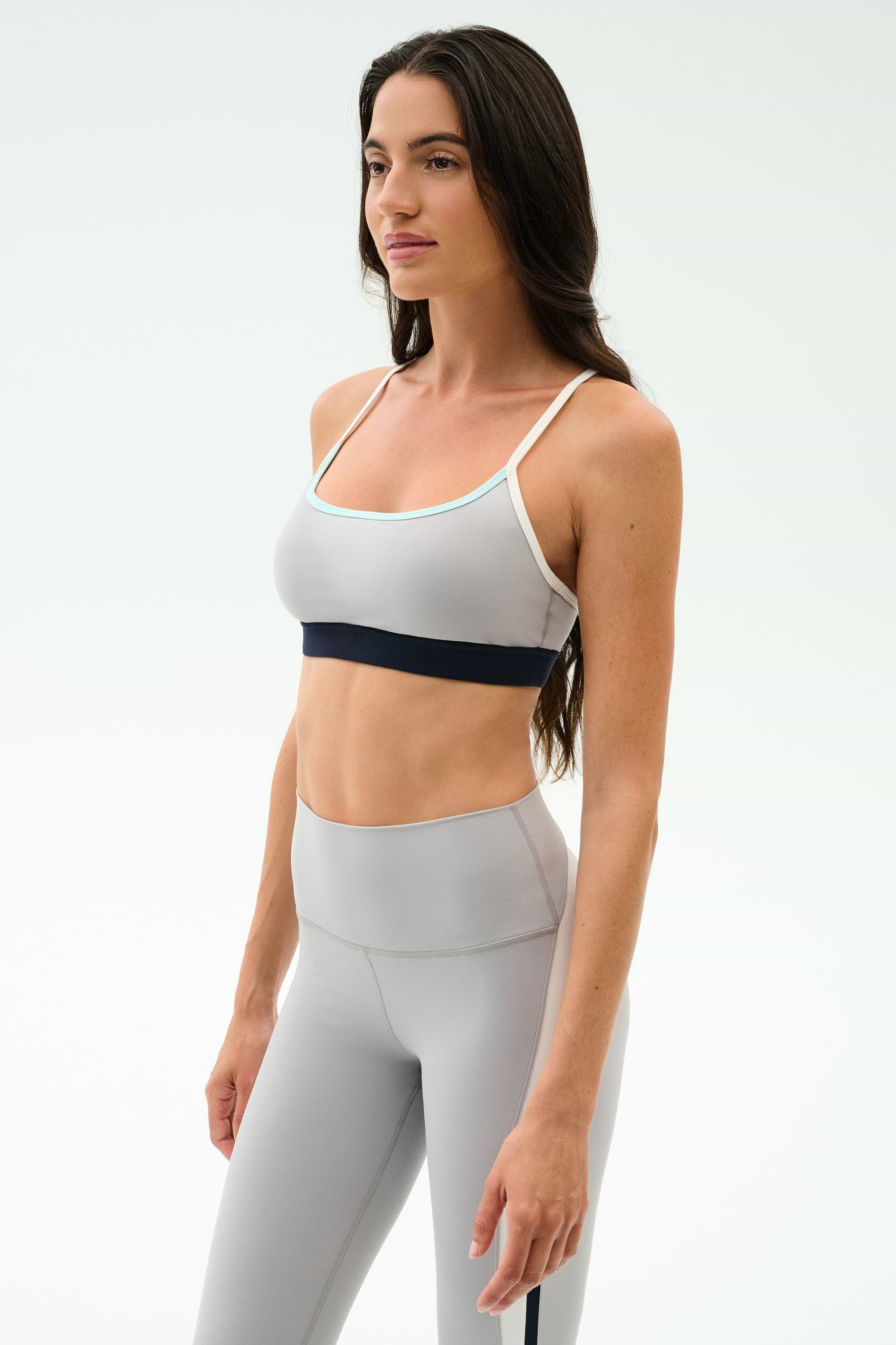 A woman in the SPLITS59 Emerson Airweight Bra in Dove Grey/Indigo and matching high-waisted leggings stands against a white background, ready for hot yoga or Pilates.