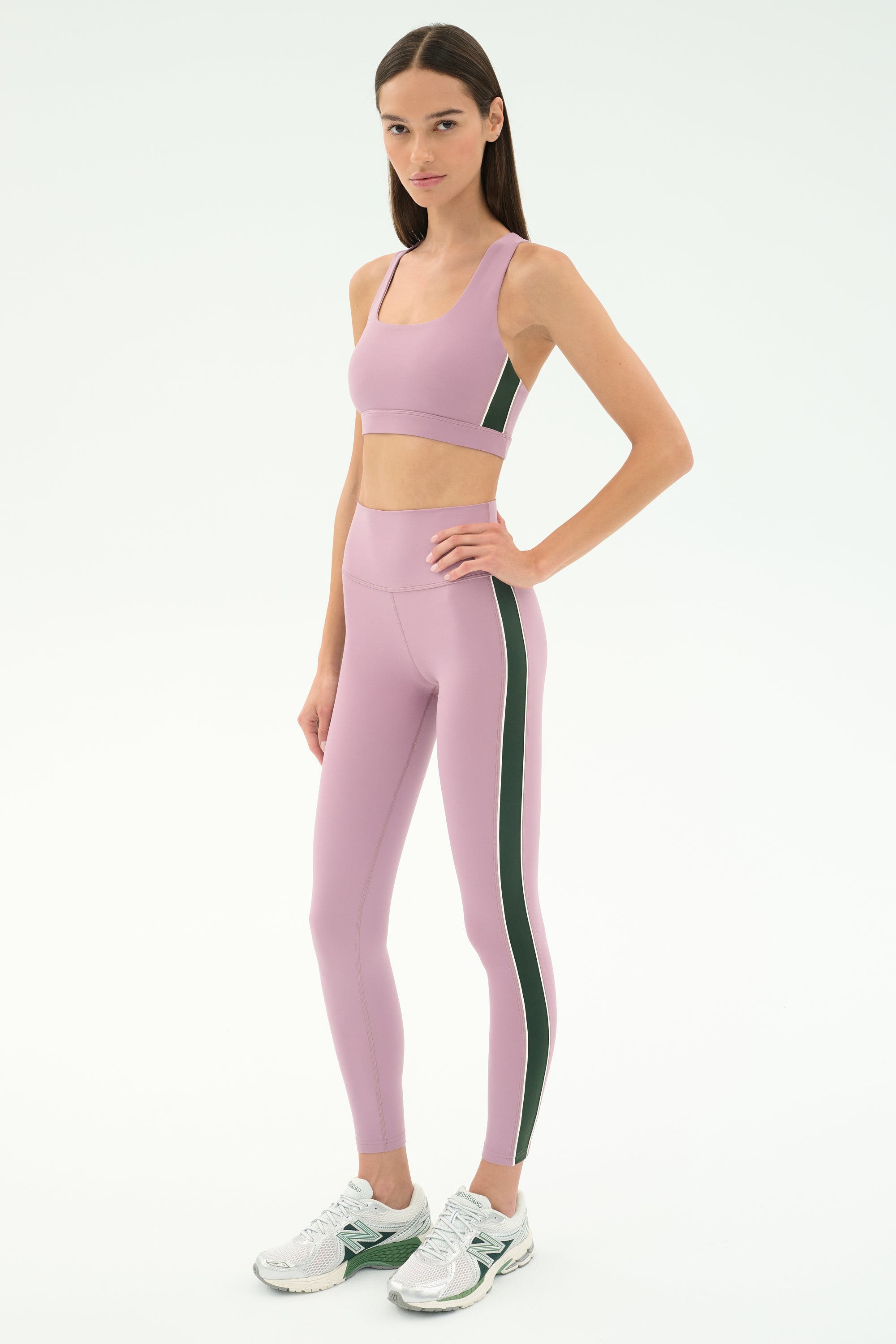 A woman stands in a studio wearing SPLITS59's Margot Rigor Legging in Blush/Military, crafted from stretchy nylon-spandex fabric, paired with a matching sports bra and white sneakers.
