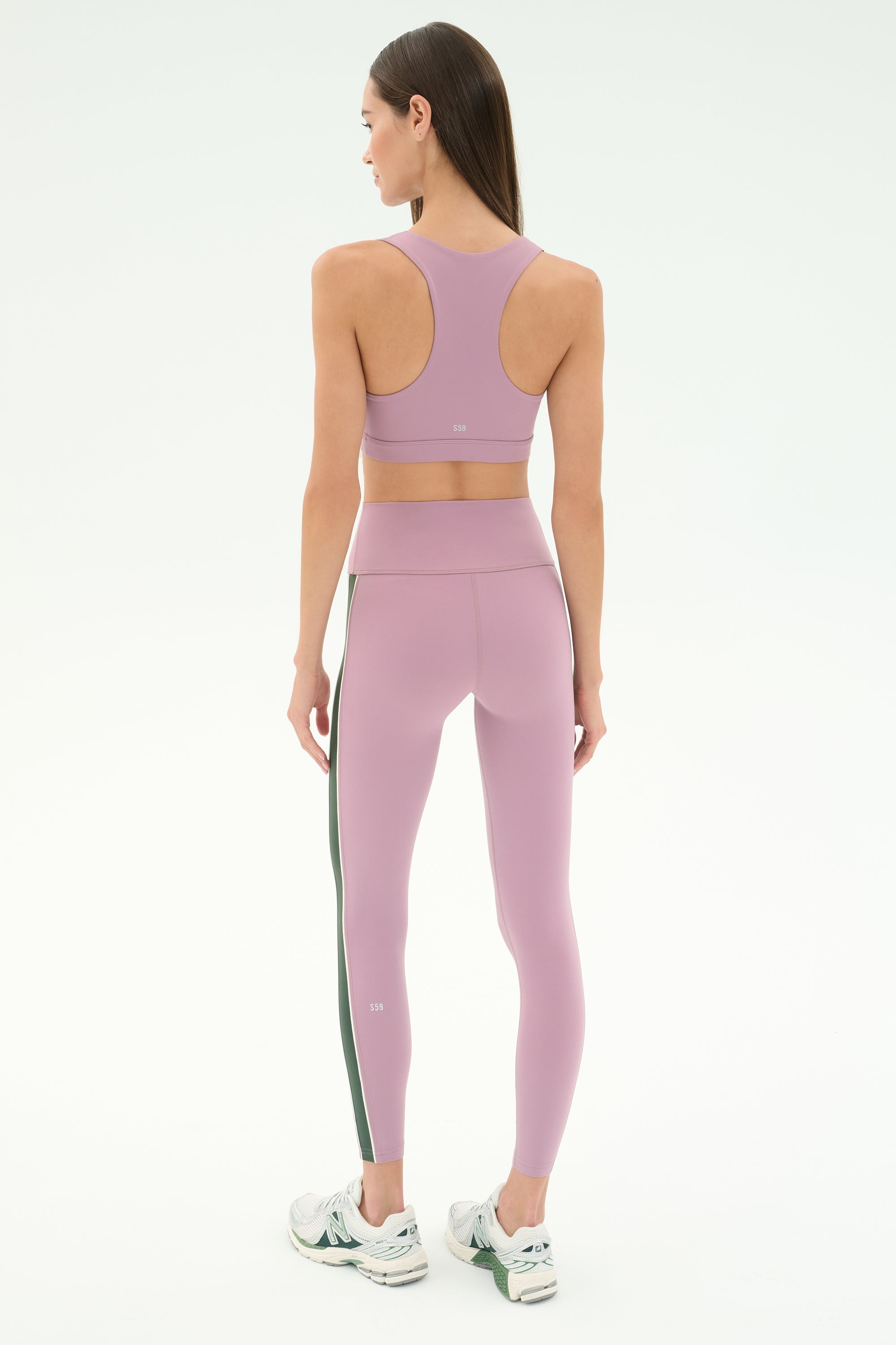 A woman faces away, wearing SPLITS59's Margot Rigor Legging in Blush/Military with a matching sports bra and white sneakers, set against a plain light background.