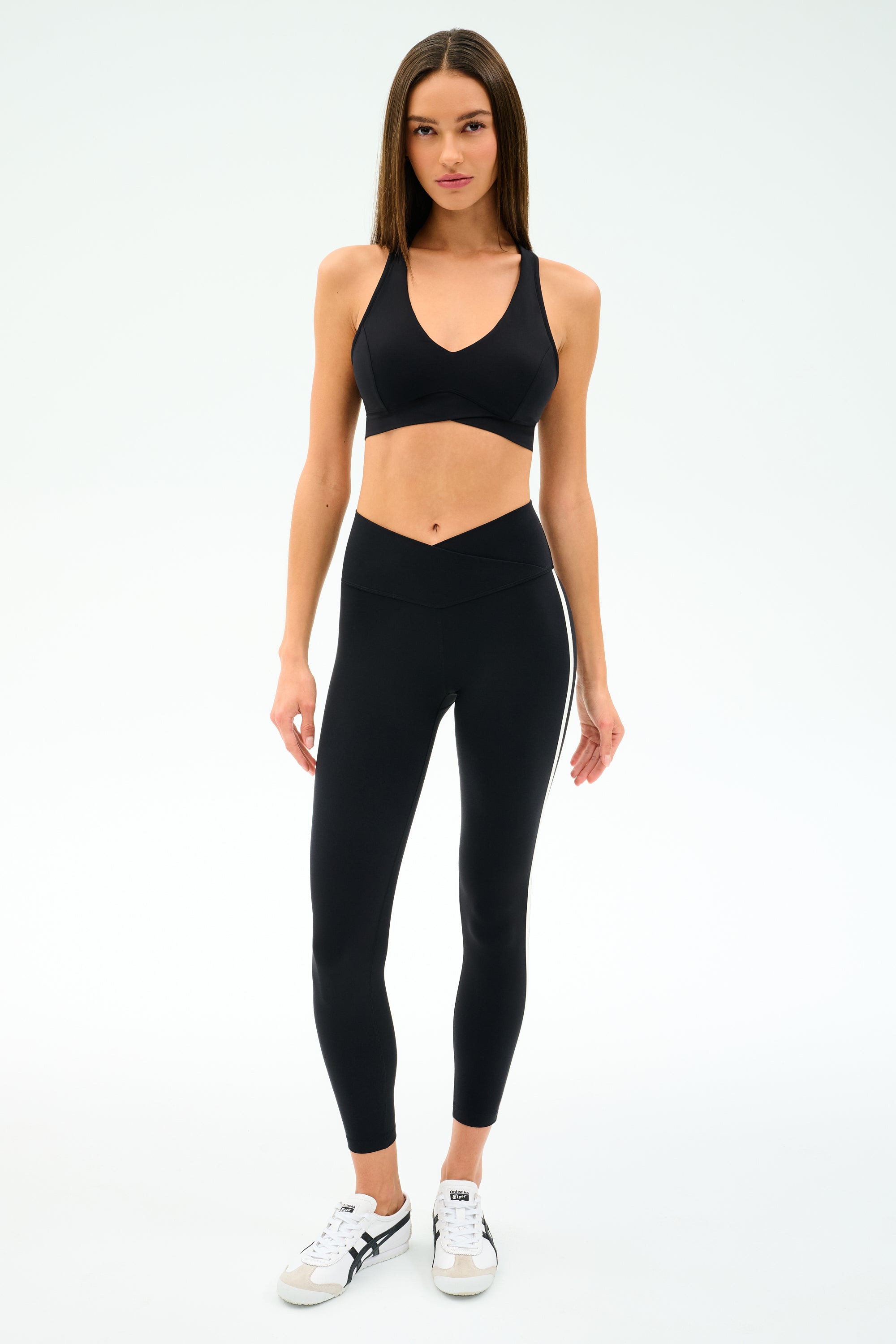 A woman faces forward wearing the SPLITS59 Mia Rigor Bra in black, paired with black high-waisted leggings and white sneakers—perfect for multi-sport performance and gym workouts—against a plain white background.