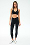 A woman faces forward against a plain white background, wearing SPLITS59’s Mia Rigor Legging w/ Stripe in Black/White, a black spandex sports bra, and white sneakers.