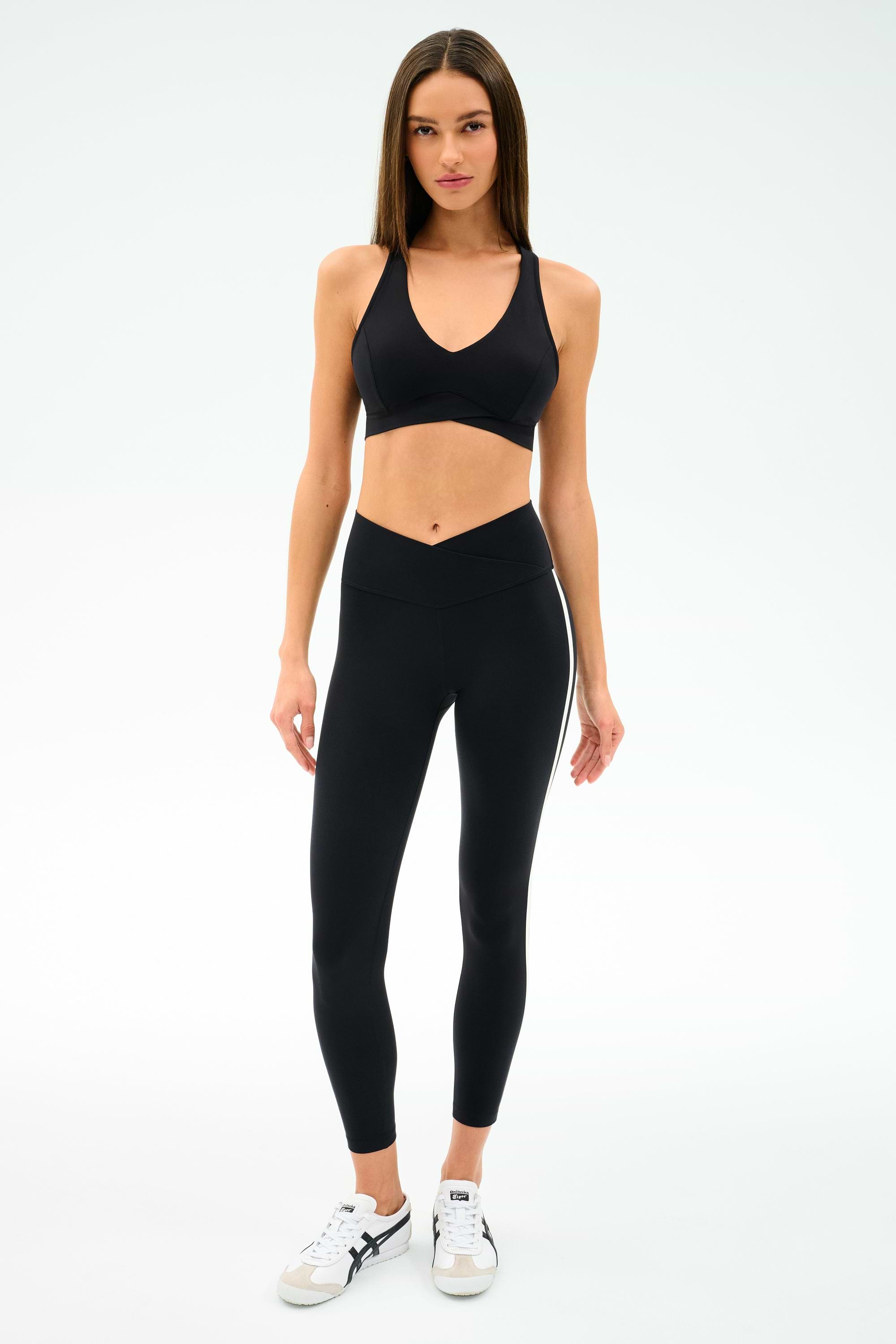 A woman faces forward against a plain white background, wearing SPLITS59’s Mia Rigor Legging w/ Stripe in Black/White, a black spandex sports bra, and white sneakers.
