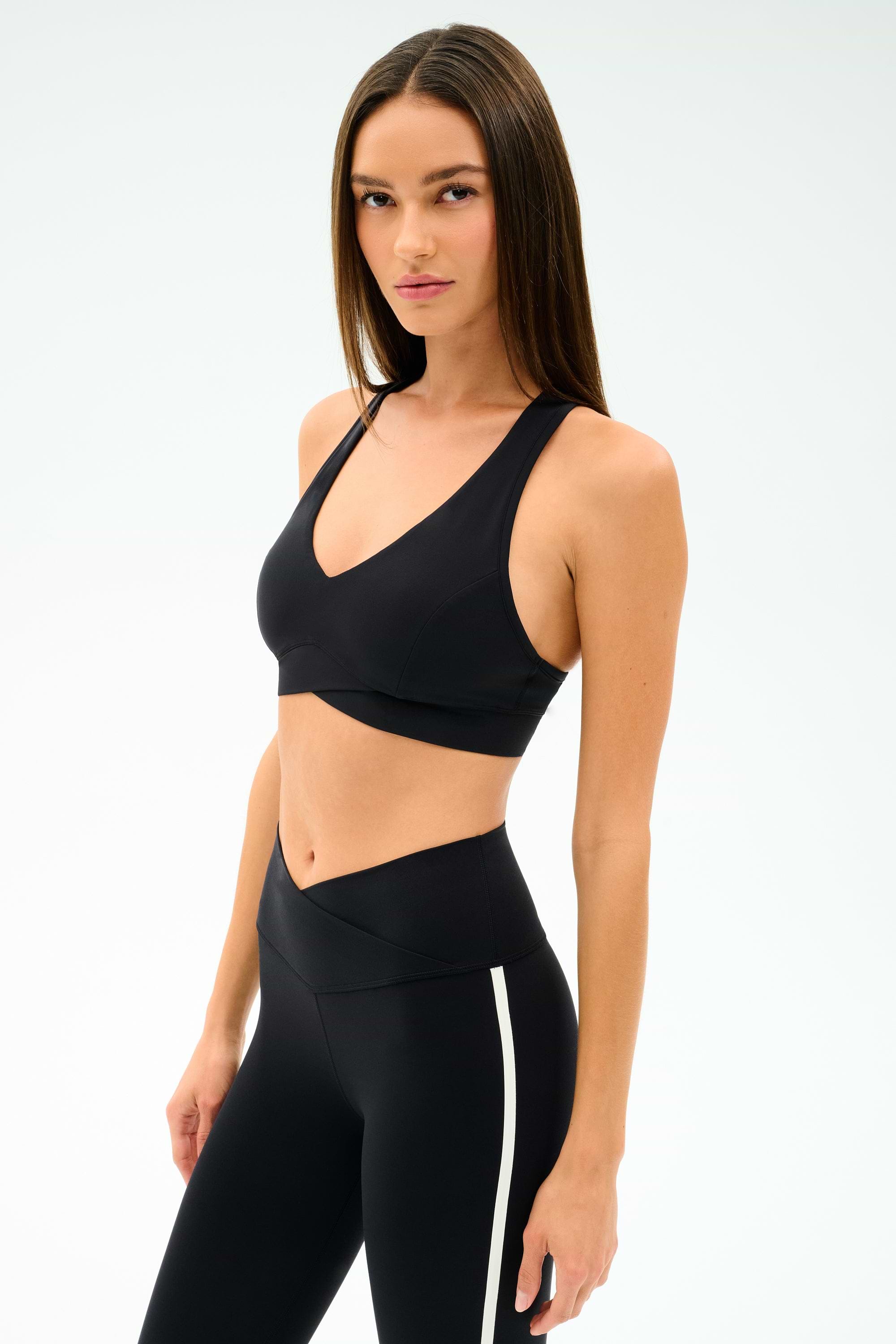 A woman models the SPLITS59 Mia Rigor Bra in black, paired with matching high-waisted leggings featuring a white stripe—ideal for multisport performance or intense gym workouts against a simple light backdrop.
