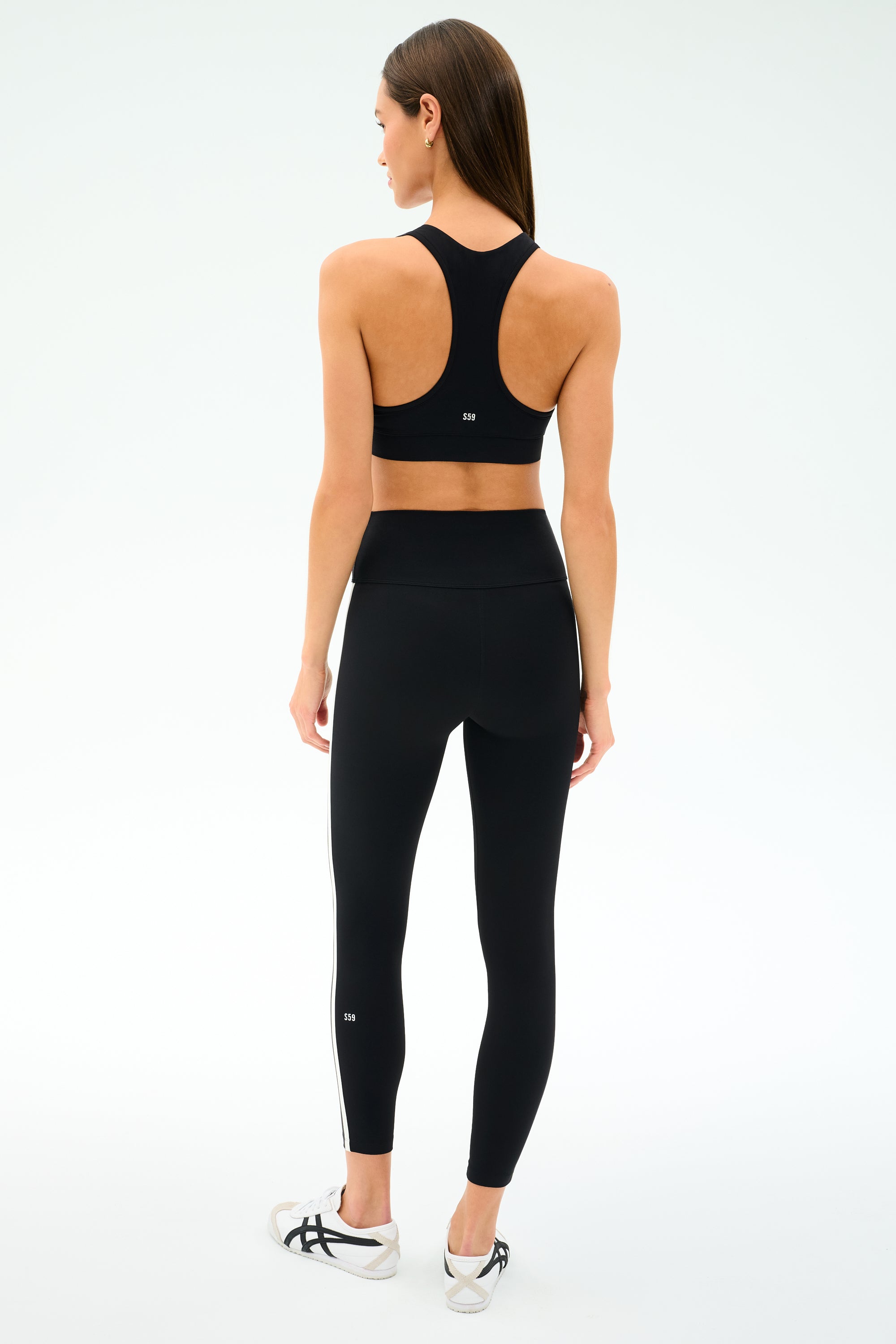 A woman stands with her back to the camera, wearing the SPLITS59 Mia Rigor Bra in black—perfect for gym workouts and multisport performance—paired with black high-waisted leggings and white sneakers against a plain white background.