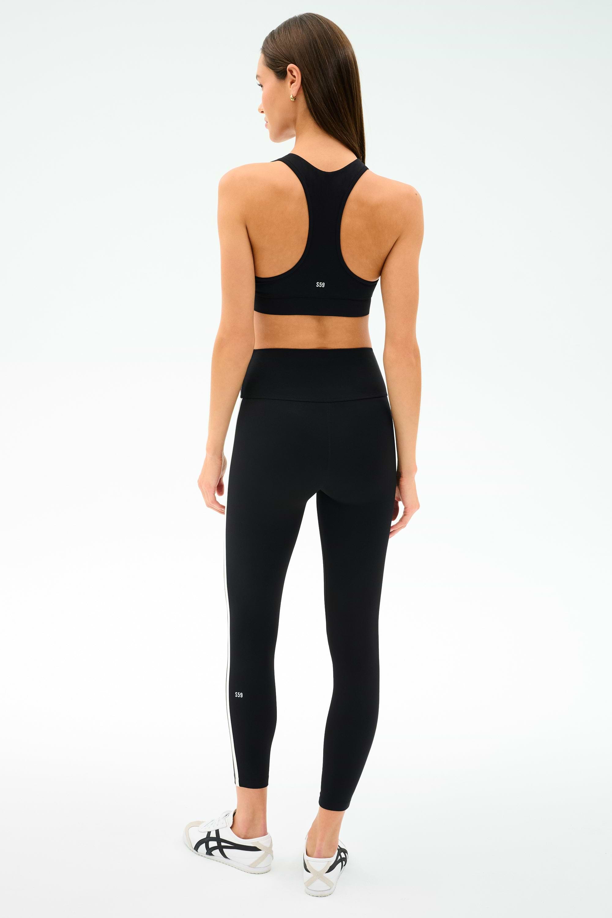 A woman stands facing away, wearing SPLITS59's Mia Rigor Legging w/ Stripe in black/white, a black nylon-spandex sports bra, and white sneakers against a plain white background.