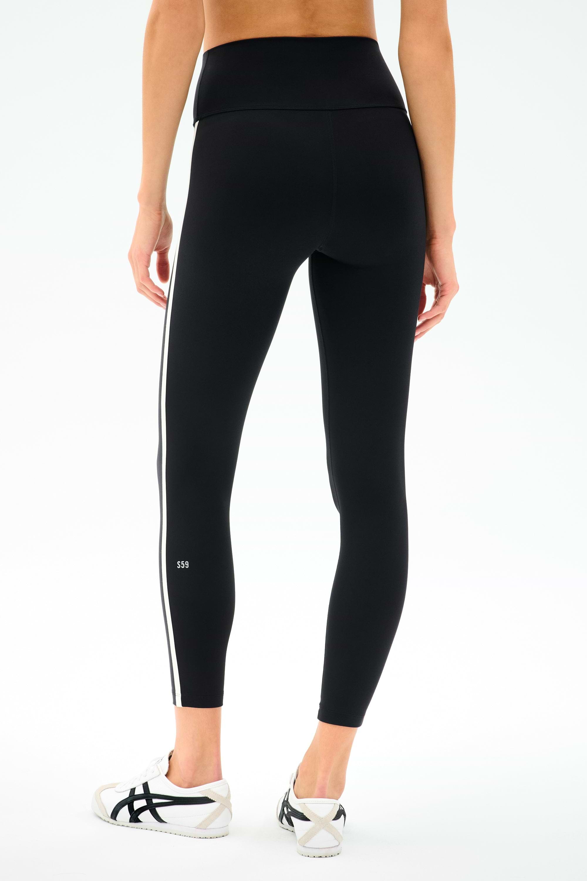A person wears the SPLITS59 Mia Rigor Legging w/ Stripe in Black/White with white athletic shoes, photographed from the back against a plain background.