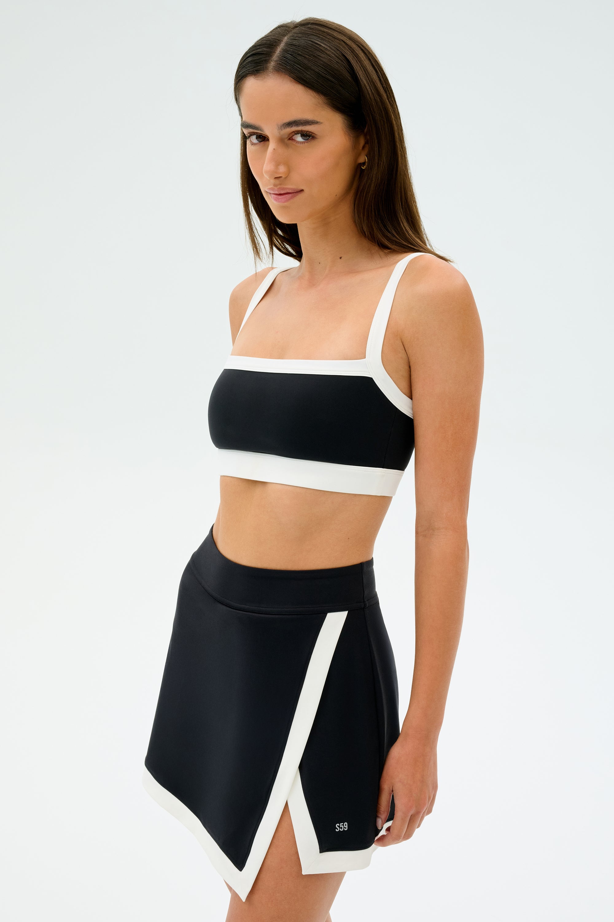 A woman confidently poses against a plain white background, ready for gym workouts, wearing the SPLITS59 Monah Rigor Bra in black and white paired with a matching skirt.