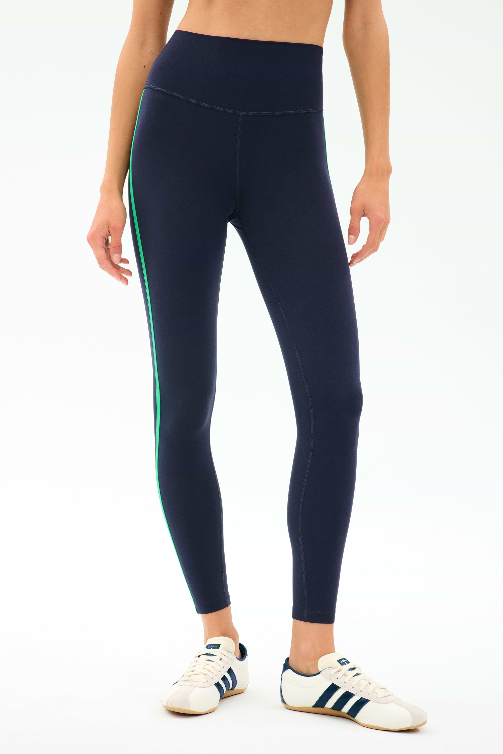 A person wearing SPLITS59 Amber Airweight High Waist 7/8 leggings in Indigo/Spearmint, featuring a green side stripe, paired with white sneakers with blue stripes, stands against a plain light background.