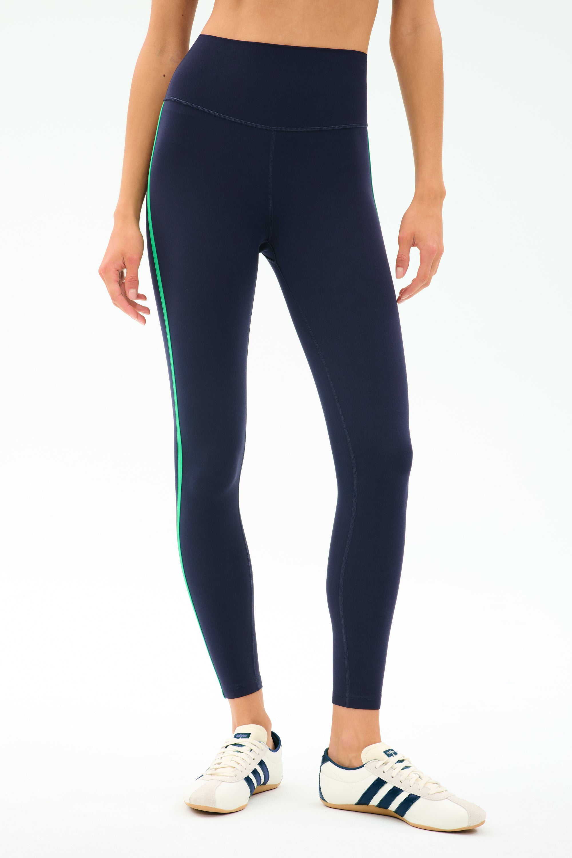 A person wearing SPLITS59 Amber Airweight High Waist 7/8 leggings in Indigo/Spearmint, featuring a green side stripe, paired with white sneakers with blue stripes, stands against a plain light background.