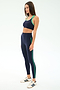 Woman wears SPLITS59 Amber Airweight High Waist 7/8 leggings in Indigo/Spearmint—navy with green trim—and white Adidas sneakers, standing against a light background and facing left in a relaxed pose.