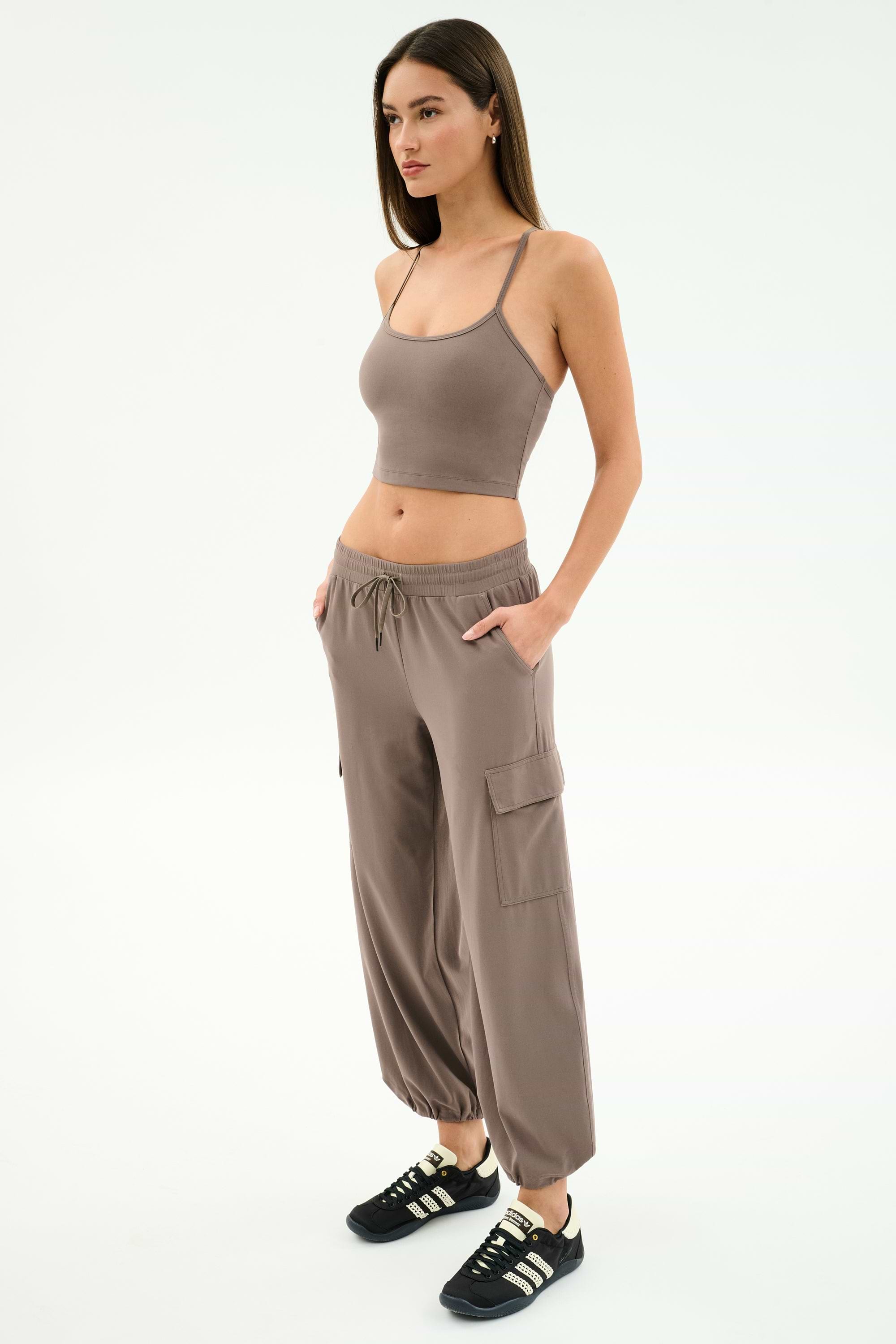 A woman poses against a white background in the SPLITS59 Airweight Crop - Lentil and matching cargo pants, paired with black sneakers with white stripes. Her hands in her pockets showcase an effortlessly cool look for hot yoga or casual outings.