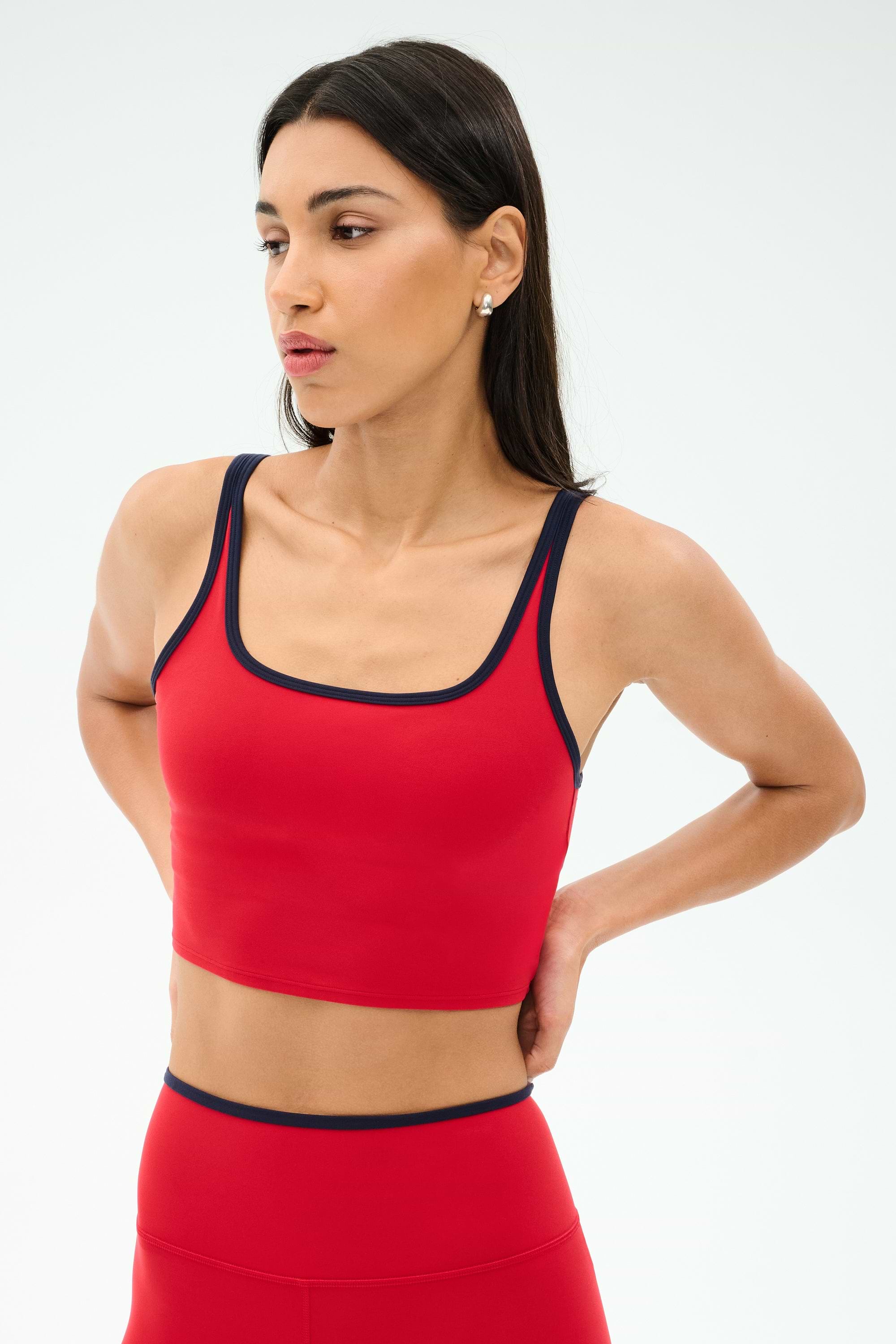 A woman models the SPLITS59 Cait Rigor Bralette in Pirate Red/Indigo, featuring black trim, with matching leggings, standing hands behind her back and looking to the side against a plain background.