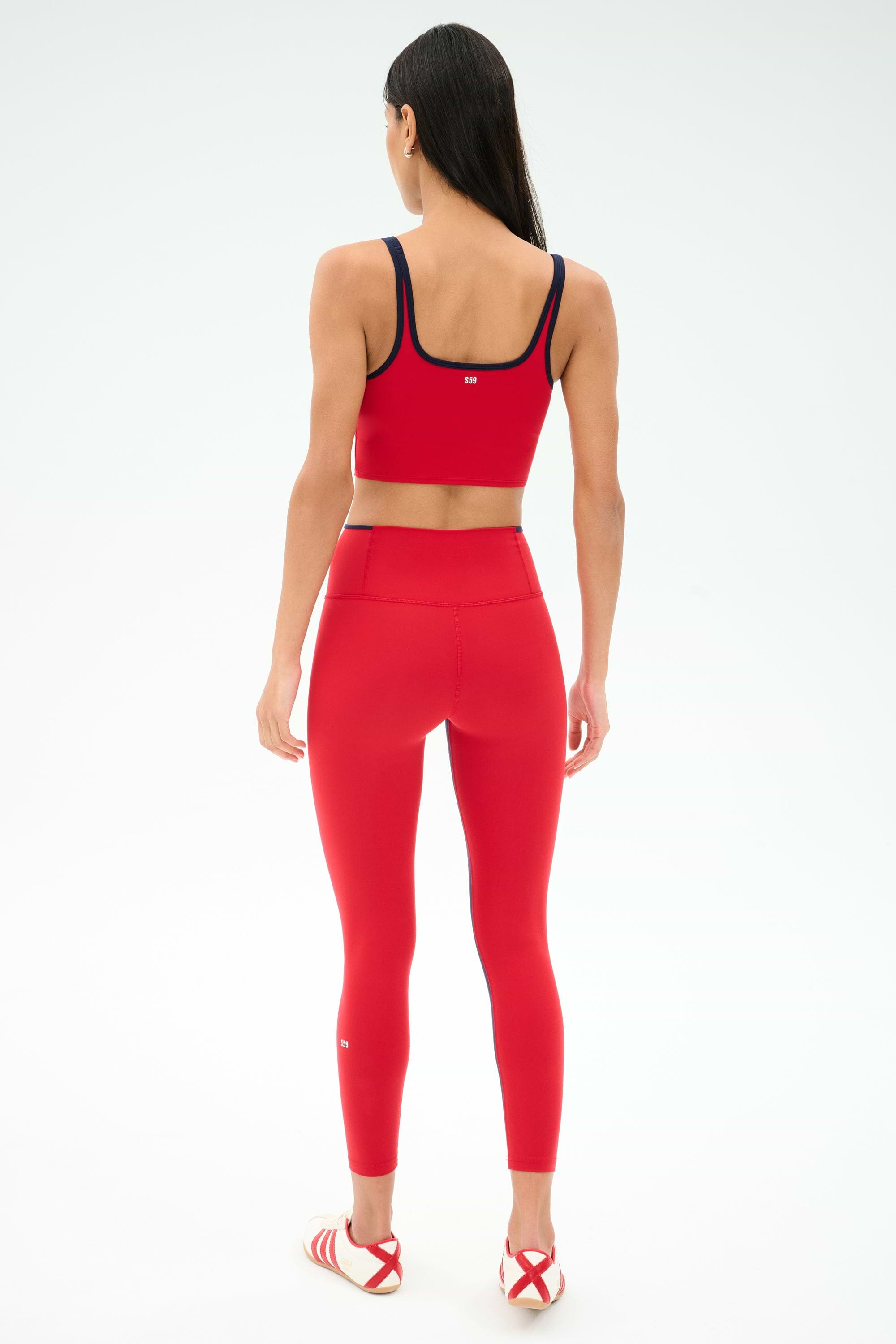 A woman faces away, wearing SPLITS59 Easton Rigor High Waist 7/8 leggings in Pirate Red/Indigo and a matching sports bra, paired with red-and-white athletic sandals against a plain light background.