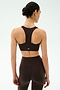 A woman with her hair in a bun faces away, wearing the SPLITS59 Sara Airweight Bra in Dark Chocolate and high-waisted leggings, ready for Pilates against a plain light background.