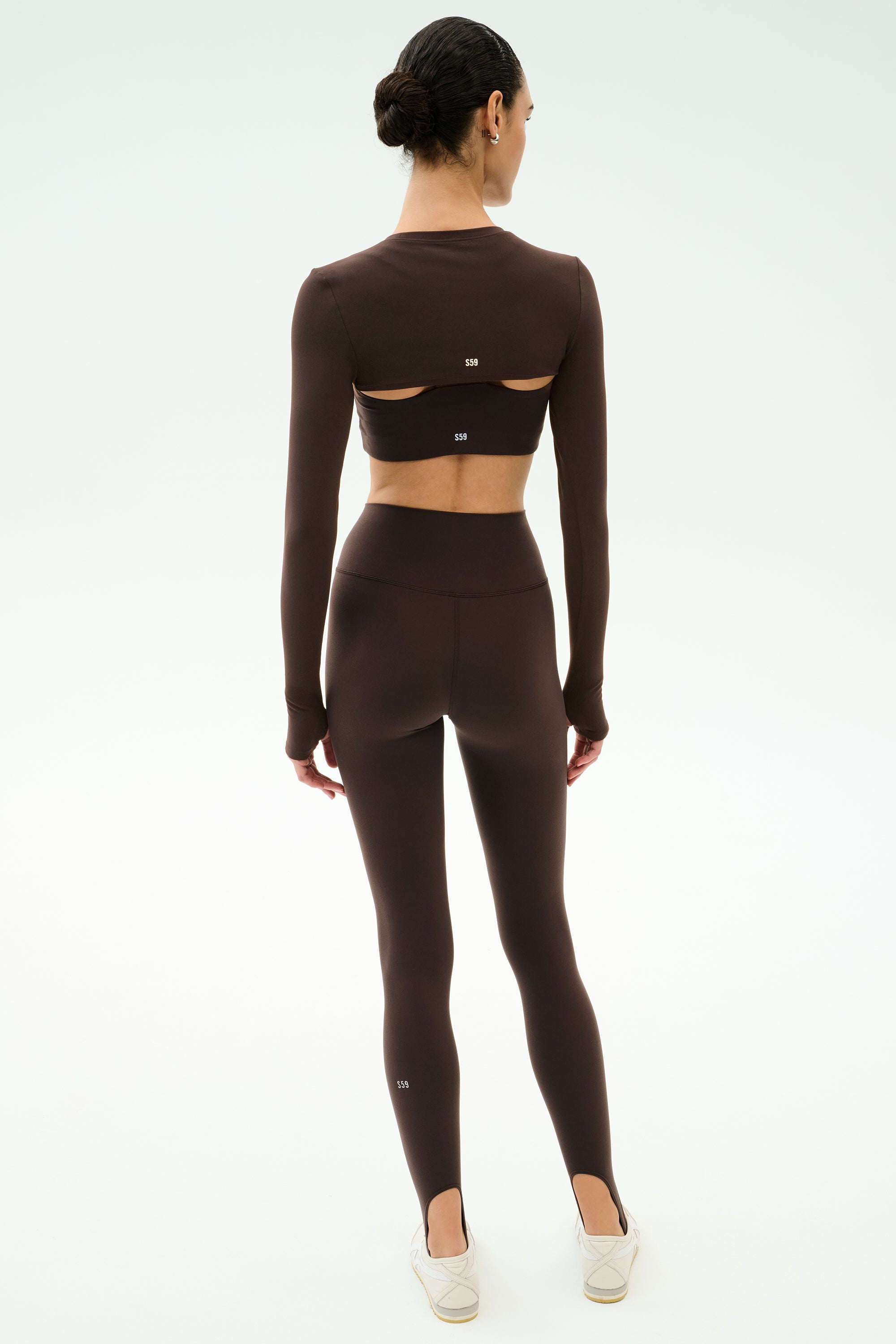 A person stands facing away, wearing the SPLITS59 Amelia Airweight Lite Shrug in Dark Chocolate with high-waisted leggings, ideal for yoga or dance, paired with white slip-on shoes against a plain light background.