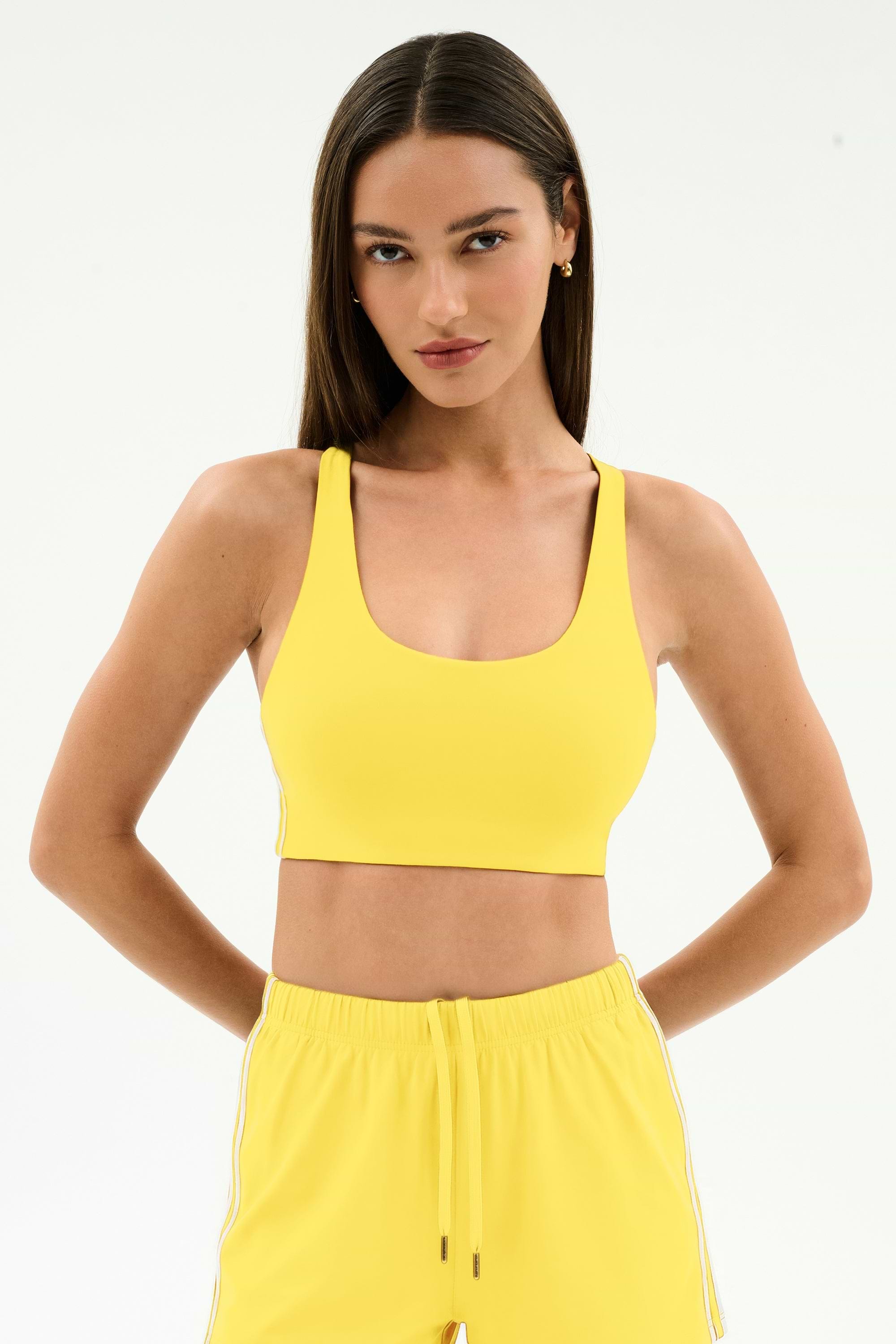 A woman wears the SPLITS59 Ella Airweight Bra in Bright Yellow/White with matching shorts, standing hands behind her back against a plain white background—ideal for hot yoga sessions.