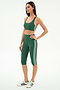 A woman stands in a studio wearing the SPLITS59 Ella High Waist Airweight Capri in Arugula/White, paired with a green sports bra and white sneakers with green stripes.