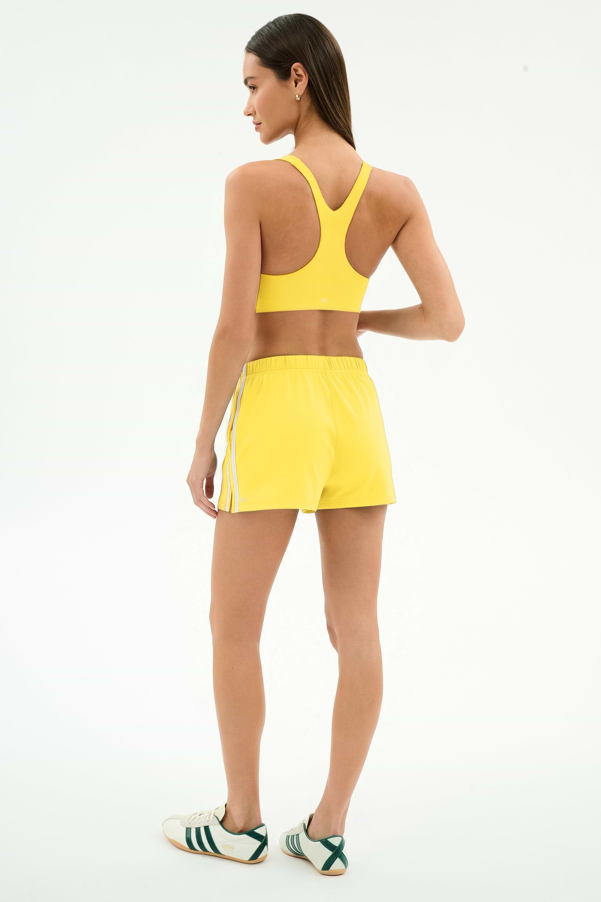 A woman stands facing away, wearing the SPLITS59 Ella Airweight Bra in Bright Yellow/White, matching yellow shorts with white stripes, and white sneakers with green stripes.