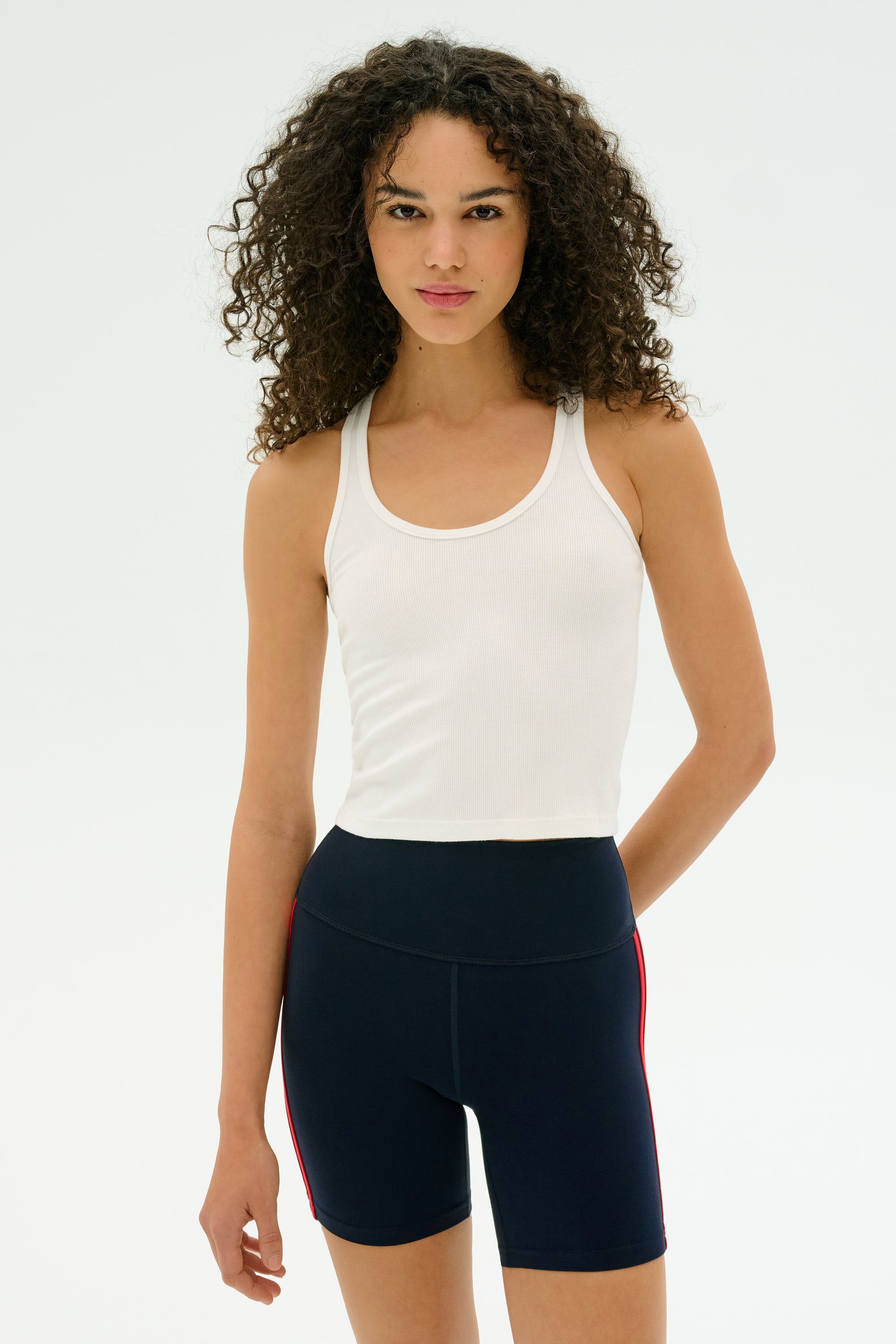 A person with curly hair wears the SPLITS59 Ashby Rib Crop tank in white and navy blue high-waisted shorts with red side stripes, standing against a plain light background.