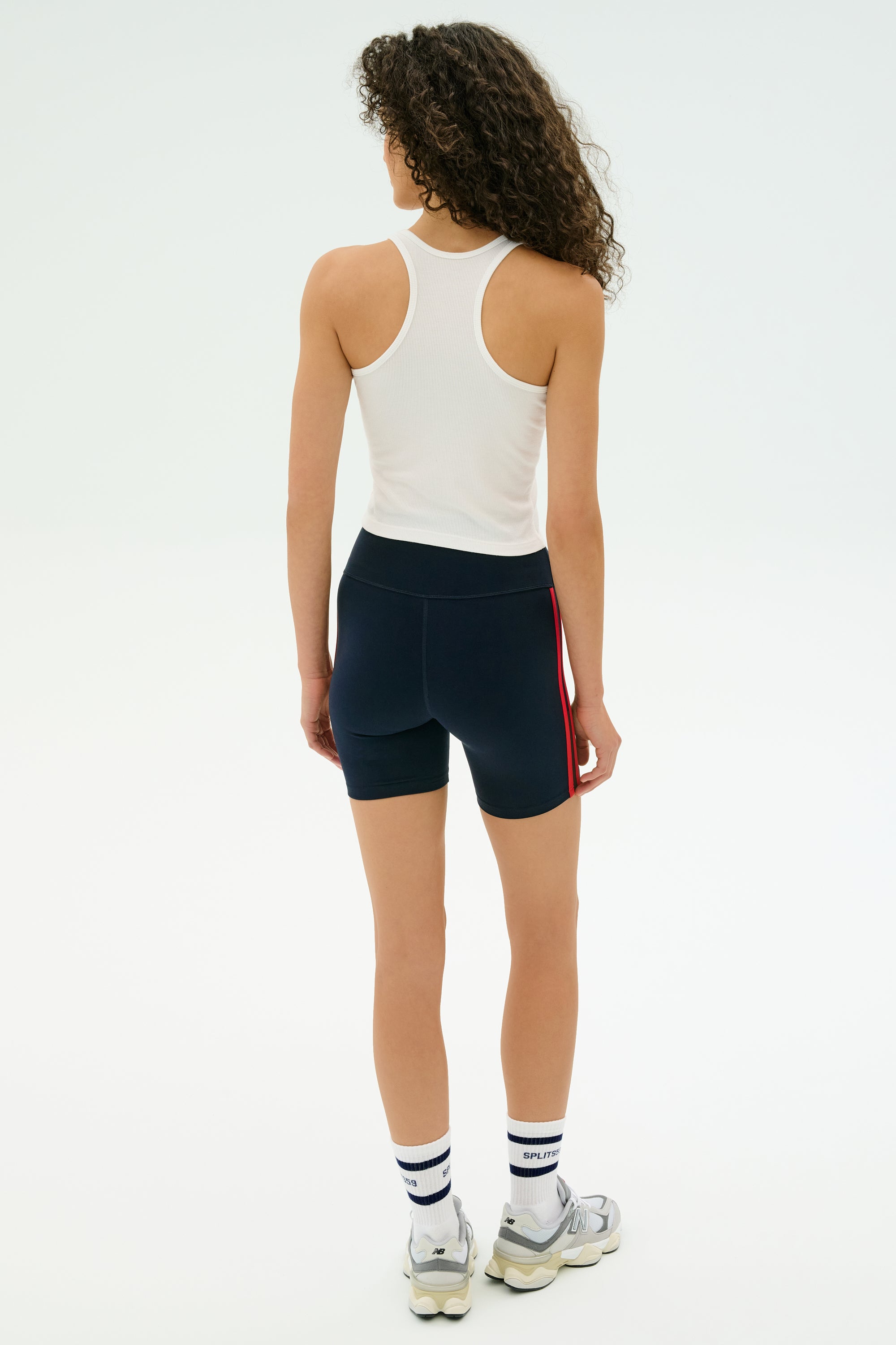 A person with curly hair, wearing the SPLITS59 Ashby Rib Crop - White, black athletic shorts with a red stripe, white socks, and sneakers stands facing away against a plain light background—ideal for gym workouts.